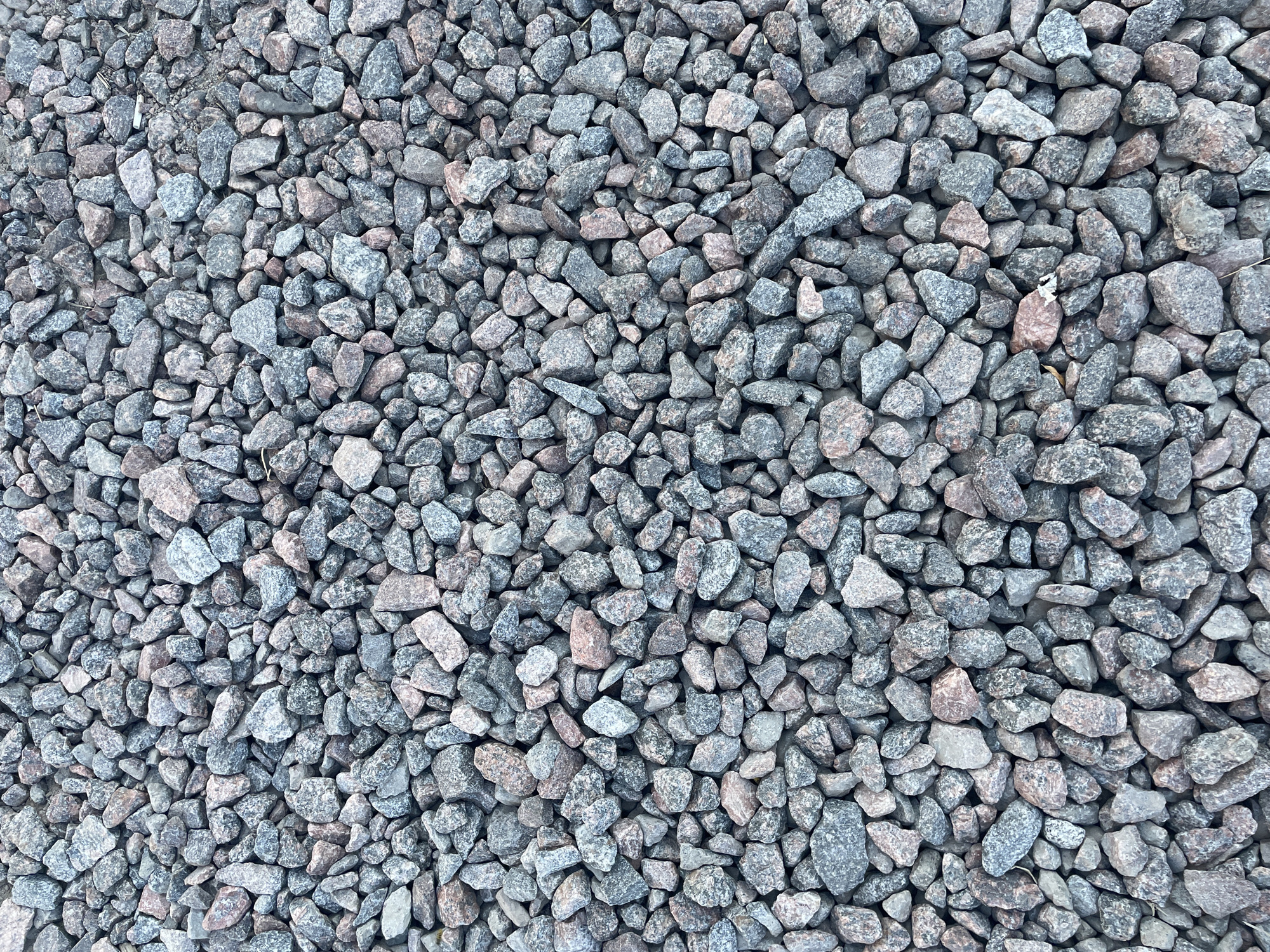 The image is a top-down view of a surface covered in coarse gravel. The gravel is composed of irregularly shaped stones, varying in size and color. Dominant colors include shades of gray, blue-gray, and some pinkish-brown hues, suggesting a mixture of granite or similar rock types. The stones are densely packed, with minimal visible spaces between them. The lighting appears diffused, casting soft shadows that highlight the texture and three-dimensionality of the gravel. There are no people, animals, or discernible man-made structures or objects in the image, creating an abstract composition of natural materials. The environment appears to be an outdoor, unpaved area. There is no information available in the image to suggest its location in the city of Unknown, Russia.