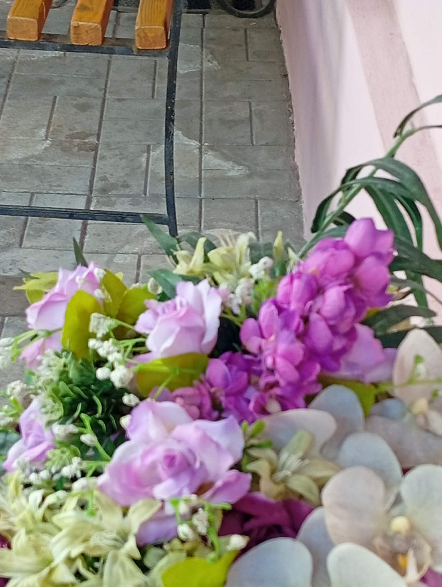The image shows a floral arrangement with pink, purple, and white flowers. Some green foliage is also visible. The flowers are arranged near a bench and a tiled walkway, positioned close to a pink wall. I have no information to confirm the location as the city of Poltava, Ukraine.