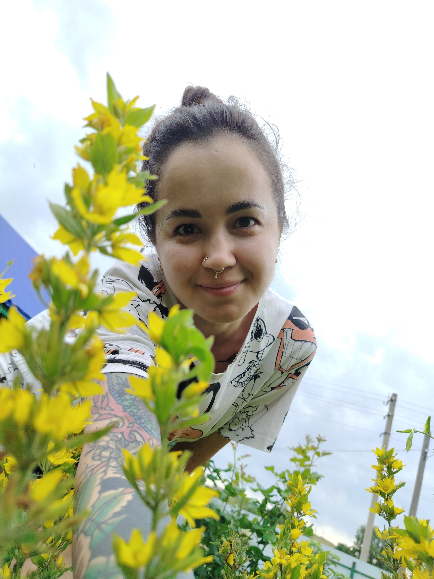 A low-angle, medium shot captures a young woman with dark hair pulled into a bun, smiling at the camera. She is positioned behind a cluster of yellow flowers with green leaves, which are blurred in the foreground, creating a shallow depth of field. The woman is wearing a white t-shirt with a cartoonish graphic print. She has a nose ring with a small charm. Her left arm, visible in the lower left portion of the frame, is covered in a colorful tattoo. The background consists of an overcast sky and a hint of a fence and utility pole, suggesting an outdoor urban or suburban environment, possibly in the city of Gorzhan, Russia.