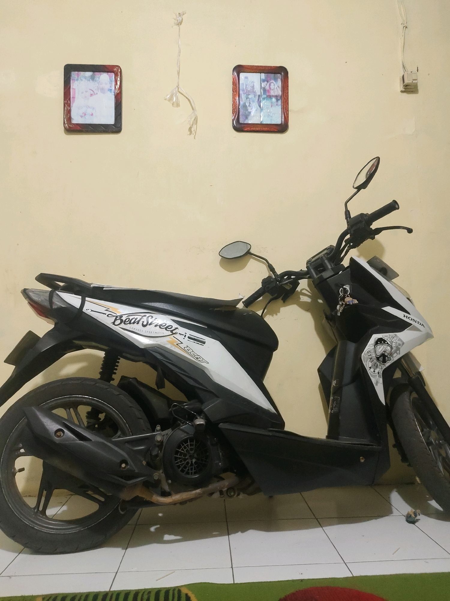 A black and white Honda Beat scooter is parked indoors against a light yellow wall. Two framed photographs are mounted on the wall above the scooter. The photograph on the left depicts a couple in wedding attire. The photograph on the right contains multiple smaller portraits. A wire with frayed ends hangs from the wall near the left photograph. Electrical wiring and a socket are visible on the wall to the right of the right photograph. The scooter is positioned such that its right side is closest to the camera. A key ring with keys hangs from the scooter's handlebars. The scooter's front wheel is on the right side of the image, and the rear wheel is on the left. The scooter has "Beat Street" and "COMPACT SPORTMATIC" decals on its body. The floor appears to be tiled, with a green and red rug partially visible at the bottom of the frame. A small toy figure is on the floor near the scooter's front wheel. The scene appears to be an indoor setting, possibly a garage or a room within a residence. The overall impression is one of a typical Indonesian home environment.