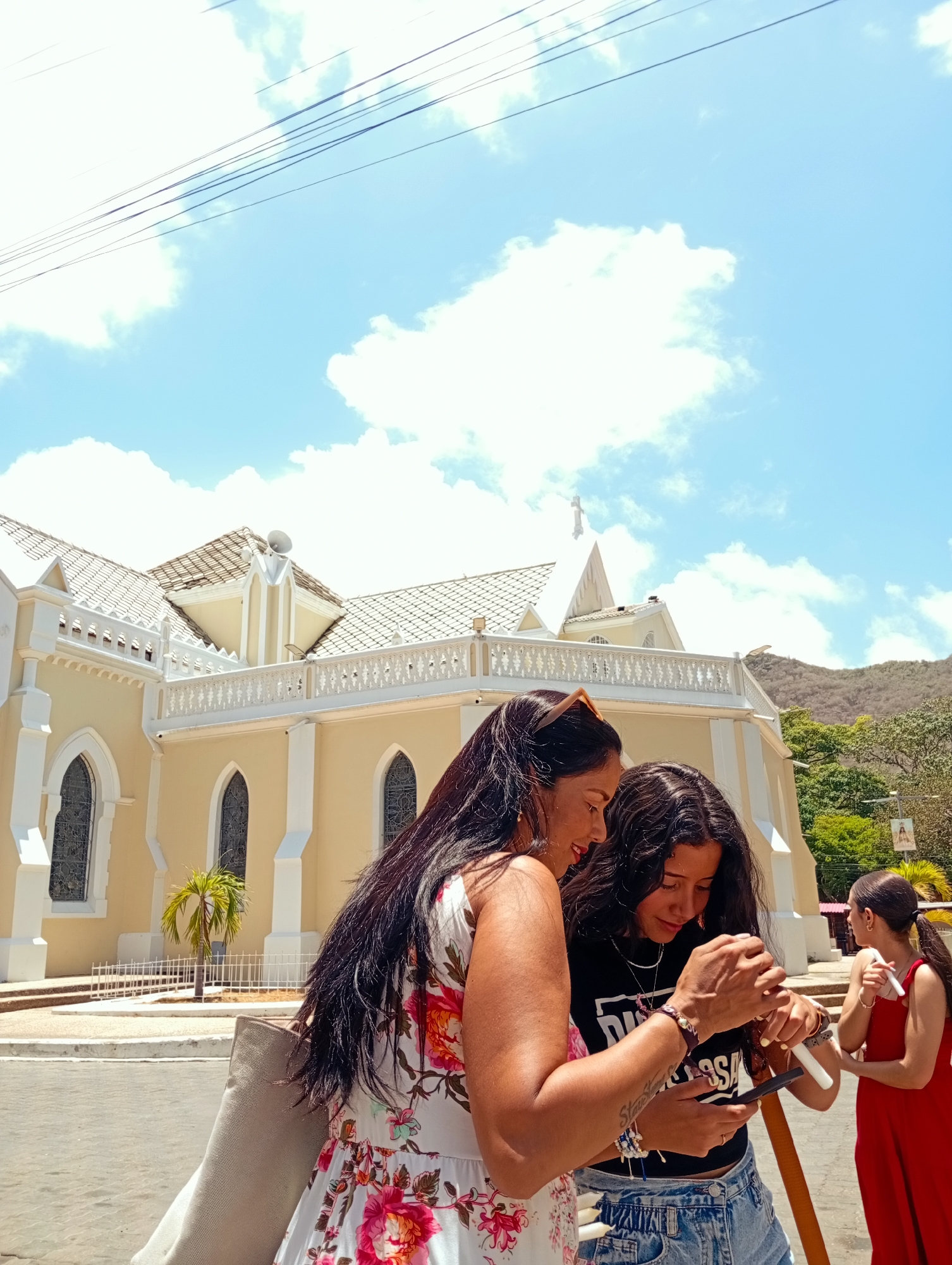 The image shows three women on a sunny day in front of the Basilica Menor de Nuestra Señora del Valle, a yellow and white church with a cross on top, located in El Valle del Espíritu Santo, Venezuela. Two of the women are standing close together and looking down at a smartphone or similar device, held by the younger of the two, with the older looking at the screen. The woman on the left is wearing a floral dress, has long dark hair, a shopping bag over her shoulder and is looking at what's happening on the cell phone. The woman next to her is wearing a black top and denim shorts. The third woman is in the background, wearing a red dress, facing away from the camera, holding a stick and tube in the left arm, while turned to the right to look at the other women. The church is behind the women, with the sky visible above it. Some power lines cross the top of the frame.