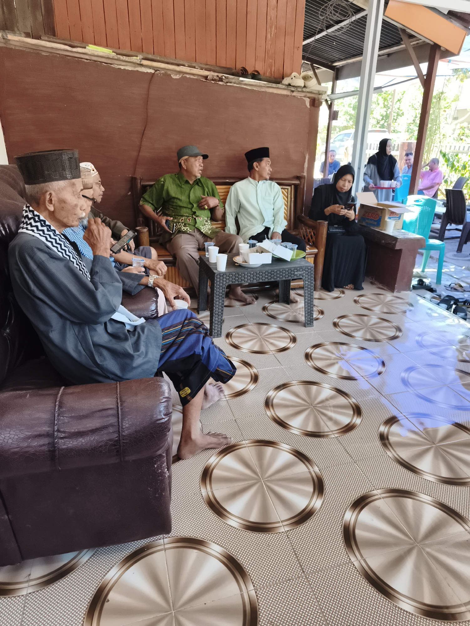In Tonasa, Indonesia, a group of people, predominantly men in traditional attire, are gathered outdoors, seated on a sofa and wooden benches. An elderly man in a gray robe and a black skullcap is seated on the left, holding a white cup and a mobile phone. Next to him, another man in a white skullcap is visible. Further back, a man in a green patterned shirt and a cap is seated, and a man in a light green shirt and black cap is also present. A woman in a black hijab is seated at a table, looking at her phone. A white box and several cups are on the table. The scene takes place on a tiled floor with circular metallic patterns, under a structure with a wooden wall and a corrugated roof. Some furniture, including a brown leather sofa and wooden chairs, are also in the frame. The background suggests a sunny day with foliage and other structures visible.