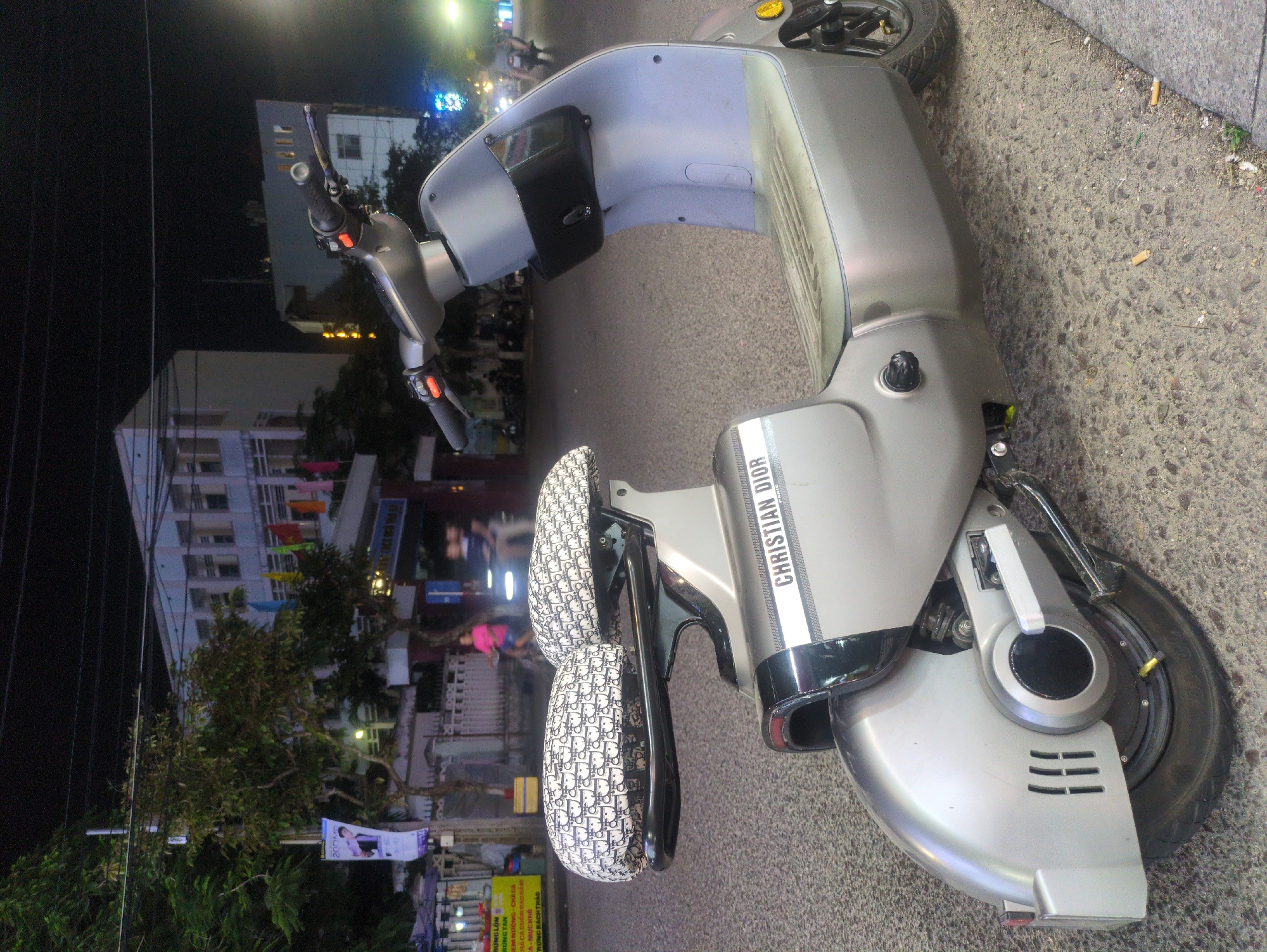 The image depicts a silver Christian Dior scooter parked on the sidewalk, presumably in South Quy Nhon Ward, Vietnam. The background includes a building adorned with colorful flags, a partially visible street sign, and a green tree. The scooter is the main subject, and its unique design is accentuated by the Christian Dior branding. The sidewalk shows signs of wear and tear. There are no visible actions or interactions.