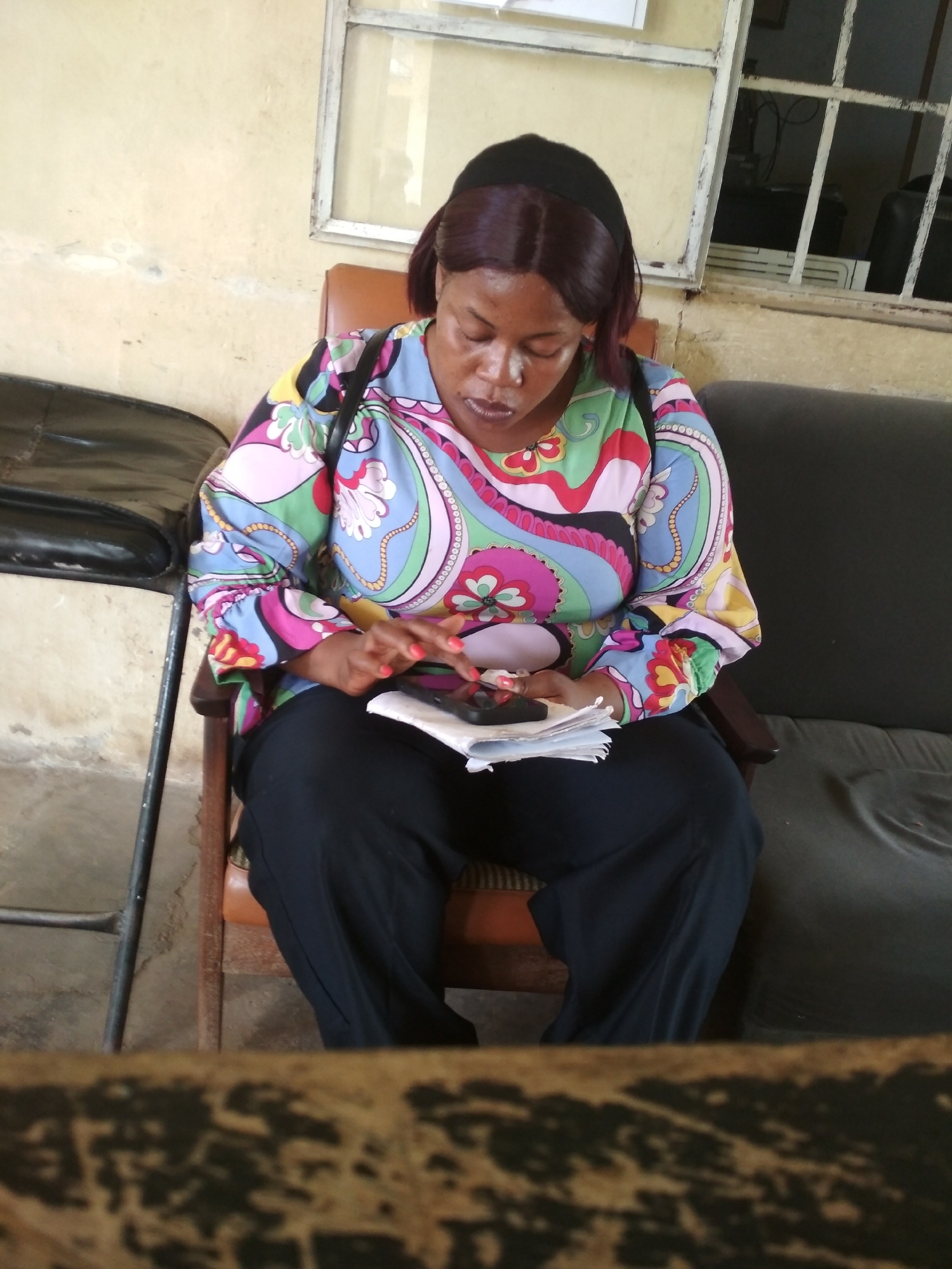 A medium shot captures a woman seated in a chair, looking down at a cell phone she holds in her hands. She is wearing a black head covering, a long-sleeved blouse with a colorful, paisley-like pattern in shades of blue, green, pink, and yellow, and dark trousers. Her fingernails are painted a bright red. She is holding a stack of papers under the phone. The woman is seated in what appears to be an office or waiting room setting. The walls are a pale yellow, and a large, barred window is visible in the background. To the left of the woman, a black chair or couch is partially visible, and to her right, a dark sofa is also in view. The floor is concrete. This scene is located in the city of Bisichi, Nigeria.