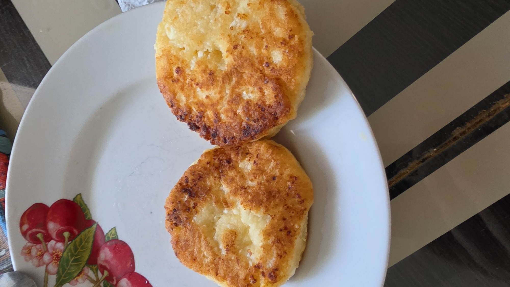 Two golden-brown, round syrniki, a type of Russian cheese pancake, are presented on a white plate with a cherry blossom design. The syrniki have a slightly uneven surface and appear to be fried to a crisp. The plate is positioned on a table with a striped surface, alternating between dark gray and beige. The background is out of focus, but hints of what might be a patterned tablecloth or placemat can be seen. There is no indication of the city of Marx, Russia, within the image itself.