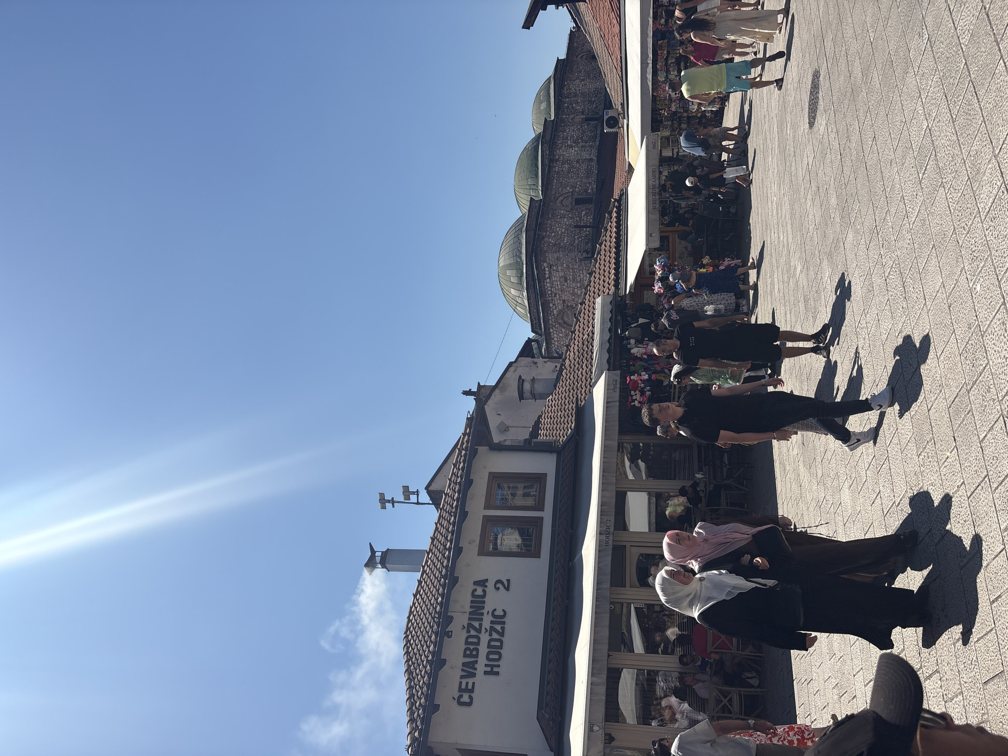 The image depicts a bustling pedestrian square in the Baščaršija district of Sarajevo, Bosnia and Herzegovina, on a clear, sunny day. Sunlight beams diagonally across the bright blue sky. In the foreground, several people are walking across a stone-paved square. Two women in hijabs, one in black and the other in a pink headscarf, walk arm-in-arm. A man in a black t-shirt and black shorts walks past them, facing away from the camera. Further in the background, more people are visible, some walking, others seated at outdoor tables.
To the left, a white building with a tiled roof features prominent signage in Bosnian Cyrillic and Latin script: "ĆEVABDŽINICA HODŽIĆ 2". This establishment appears to be a restaurant or cafe, with awnings extending over a seating area. Further down the street, several market stalls with white awnings display merchandise. In the background, the distinctive domed roofs of historic stone buildings, likely part of a mosque complex, are visible against the sky. The overall scene captures the vibrant atmosphere of a popular tourist and local gathering place in Sarajevo.