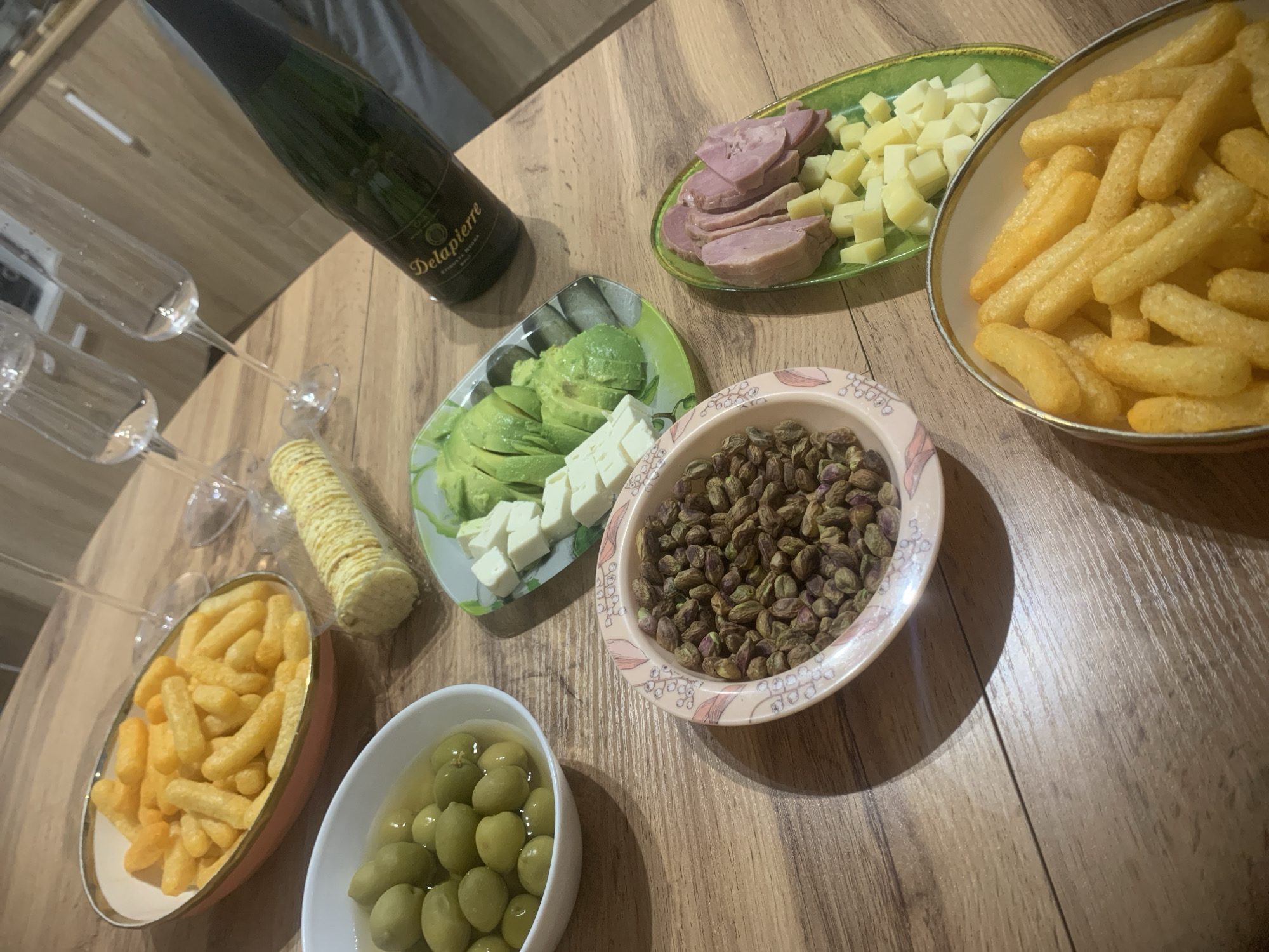 On a wood-patterned surface are several snacks and drinks. There's a bottle of Delapierre champagne, four champagne flutes, and a roll of crackers. Various plates and bowls hold sliced avocado and cheese, sliced ham and cheese cubes, puffed corn snacks, green olives, and pistachios. The location is not specified in the image.