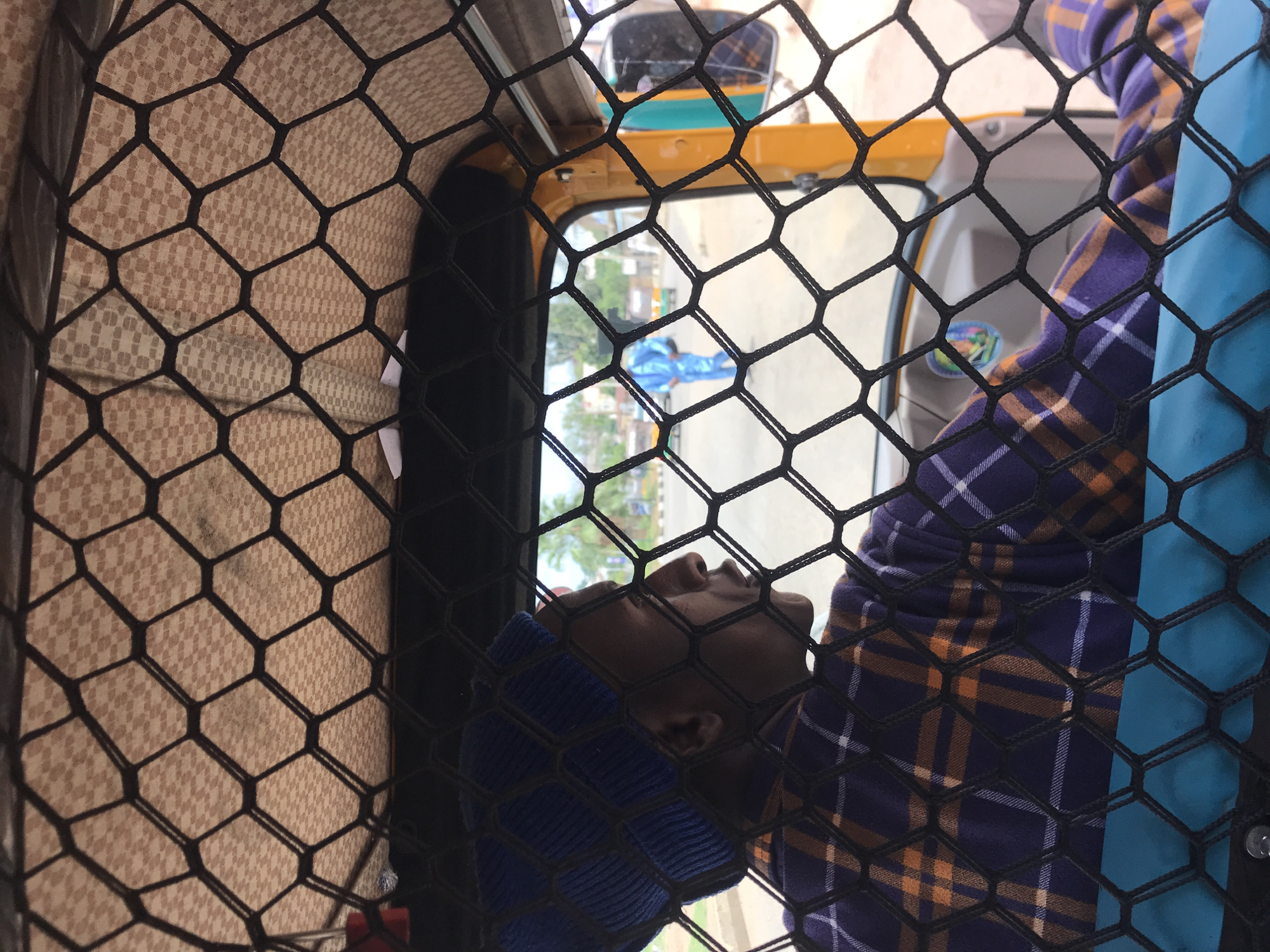 This image provides an interior perspective from a motorized three-wheeled vehicle, located in Rafin Par, Nigeria. The entire scene is viewed through a black hexagonal mesh net positioned in the immediate foreground. The vehicle's ceiling, covered in a light brown textile featuring a subtle, repeating checkered pattern, is visible at the top of the frame. On the left, the driver, a person with dark skin, is seated, facing right, and is wearing a blue knitted cap and a plaid shirt with purple, orange, and white checks. A portion of a teal-colored seat or interior panel is visible beneath the driver. Beyond the vehicle's interior and the netting, an outdoor street scene is visible. A paved road recedes into the distance, occupied by a pedestrian in a bright blue jumpsuit in the mid-ground, other vehicles, and distant buildings interspersed with vegetation, including a visible palm tree. A yellow structural component of the vehicle and its right-side mirror, reflecting a blurred exterior view, are visible on the far right.