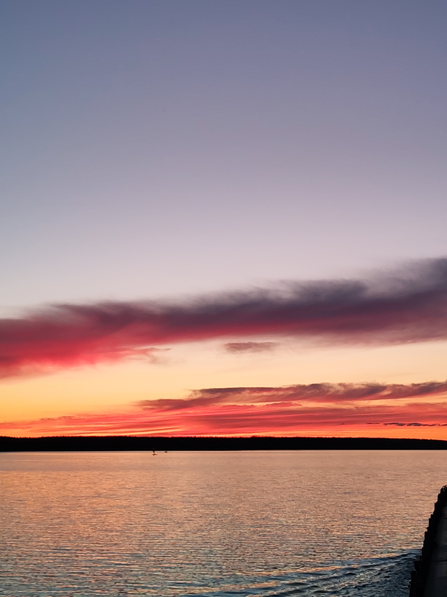 The image depicts a sunset over a body of water, likely a lake or large river. The sky transitions from a pale lavender at the top to vibrant oranges and reds near the horizon. A dark band of trees is visible on the horizon line. The water reflects the colors of the sky. There are two figures standing or floating on the water. The location is unspecified but may be Topornya, Russia.