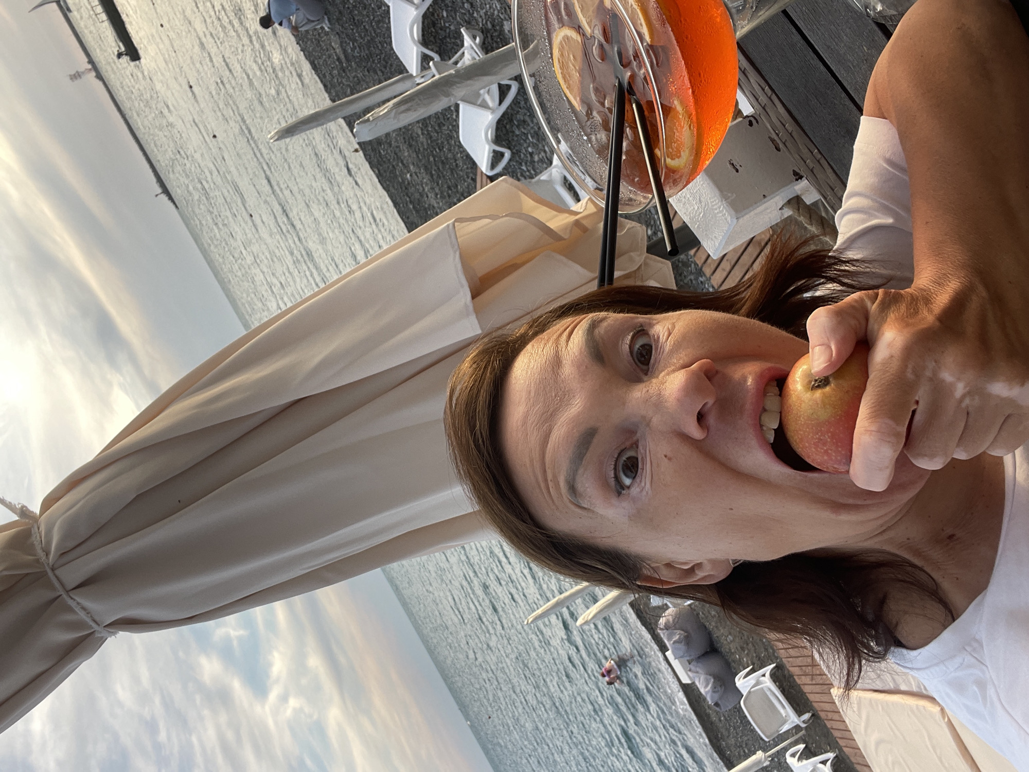 A woman with brown hair and wide eyes is taking a bite out of a red apple while holding it with her right hand. She is wearing a white t-shirt. Next to her is a large glass filled with an orange beverage, ice, and orange slices. The background shows the sea with gentle waves and a beach with lounge chairs and umbrellas. The sky is cloudy with hints of sunset colors. The scene is likely at a beachside cafe or resort in Sochi, Russia.