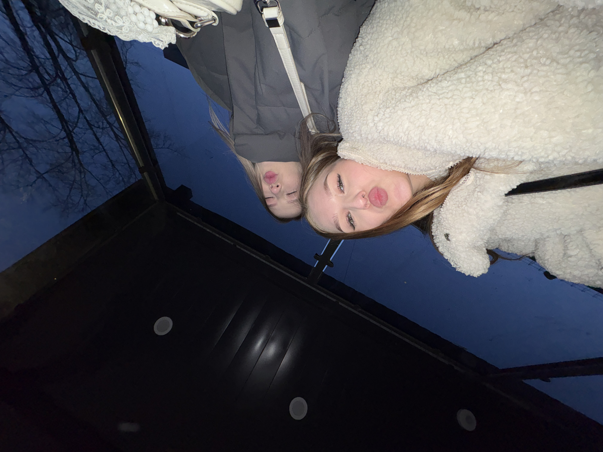 This image captures a reflective selfie of two young women, likely taken at dusk or night, in Chaikovsky, Russia. Both women are posing with pursed lips, looking into the reflective surface. The woman on the left has blonde hair, with her eyes closed, and wears a grey puffer jacket with a white shoulder bag visible. The woman on the right has light brown hair, with her eyes open, and wears a cream-colored, fluffy jacket.
The reflection reveals a dark blue sky and the silhouettes of bare tree branches in the upper left, indicating an outdoor environment beyond the reflective surface. The lower part of the reflection shows a dark, structured surface, possibly a ceiling or awning, with circular recessed lights or vents. This suggests the photo was taken from an indoor or covered outdoor area, looking up into a reflective glass surface or mirror. The overall mood is casual and playful.