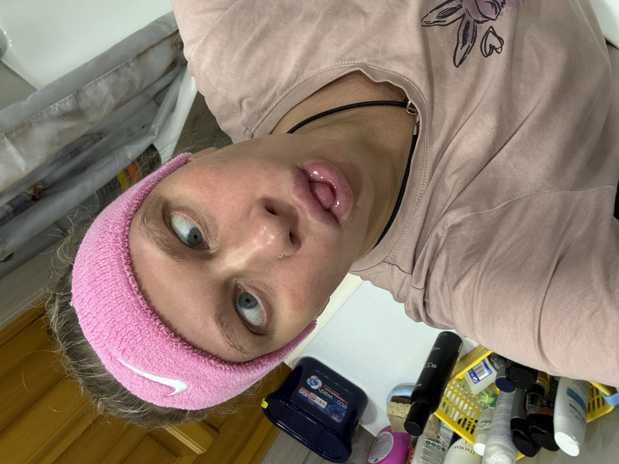 A selfie of a young woman with light skin, blue eyes, and a pink Nike headband is taken at an unusual angle, presumably indoors. Her mouth is slightly open, showing her tongue. She is wearing a light pink t-shirt with a dark design on the upper right side and a dark cord necklace. The background includes a cluttered bathroom counter with various bottles, containers, and a yellow basket. Part of a bathtub is visible on the left. The image does not provide information about the city of Perm, Russia.
