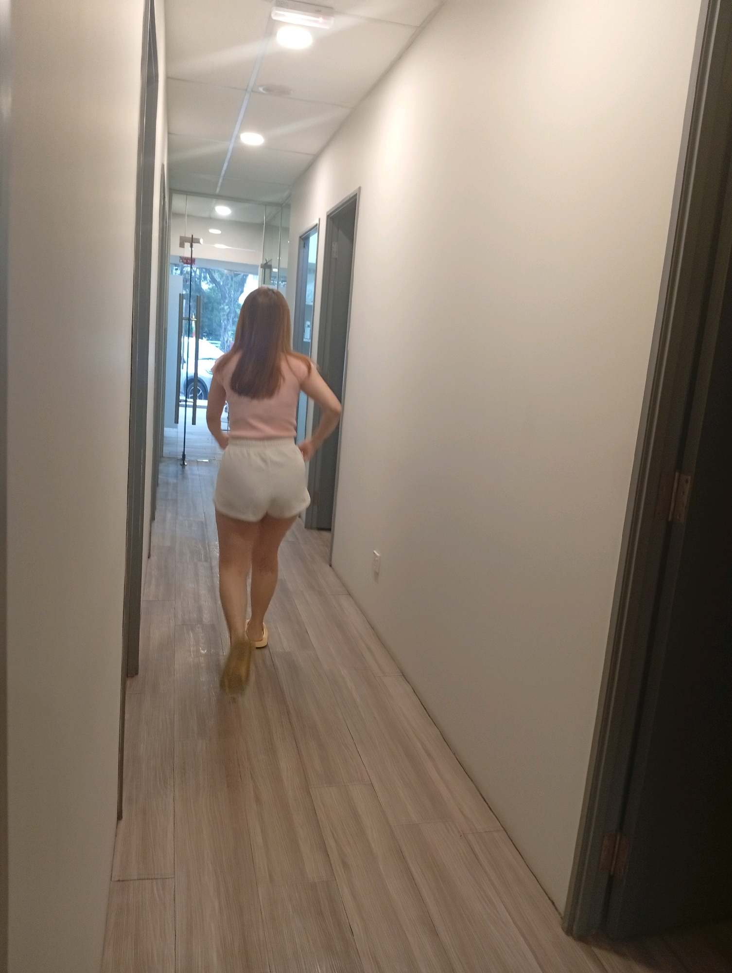 A woman with long, auburn hair is walking down a hallway. She is wearing a light pink, short-sleeved top and white shorts. She is also wearing light-colored shoes. The hallway has white walls and a light wood-look tiled floor. There are several doors on the right side of the hallway. At the end of the hallway, there is a glass door that leads outside, and a car is visible. The image appears to have been taken in a commercial building, possibly an office or a spa. The overall lighting is bright and appears to be artificial. The location is in the Selayang Municipal Council, Malaysia.