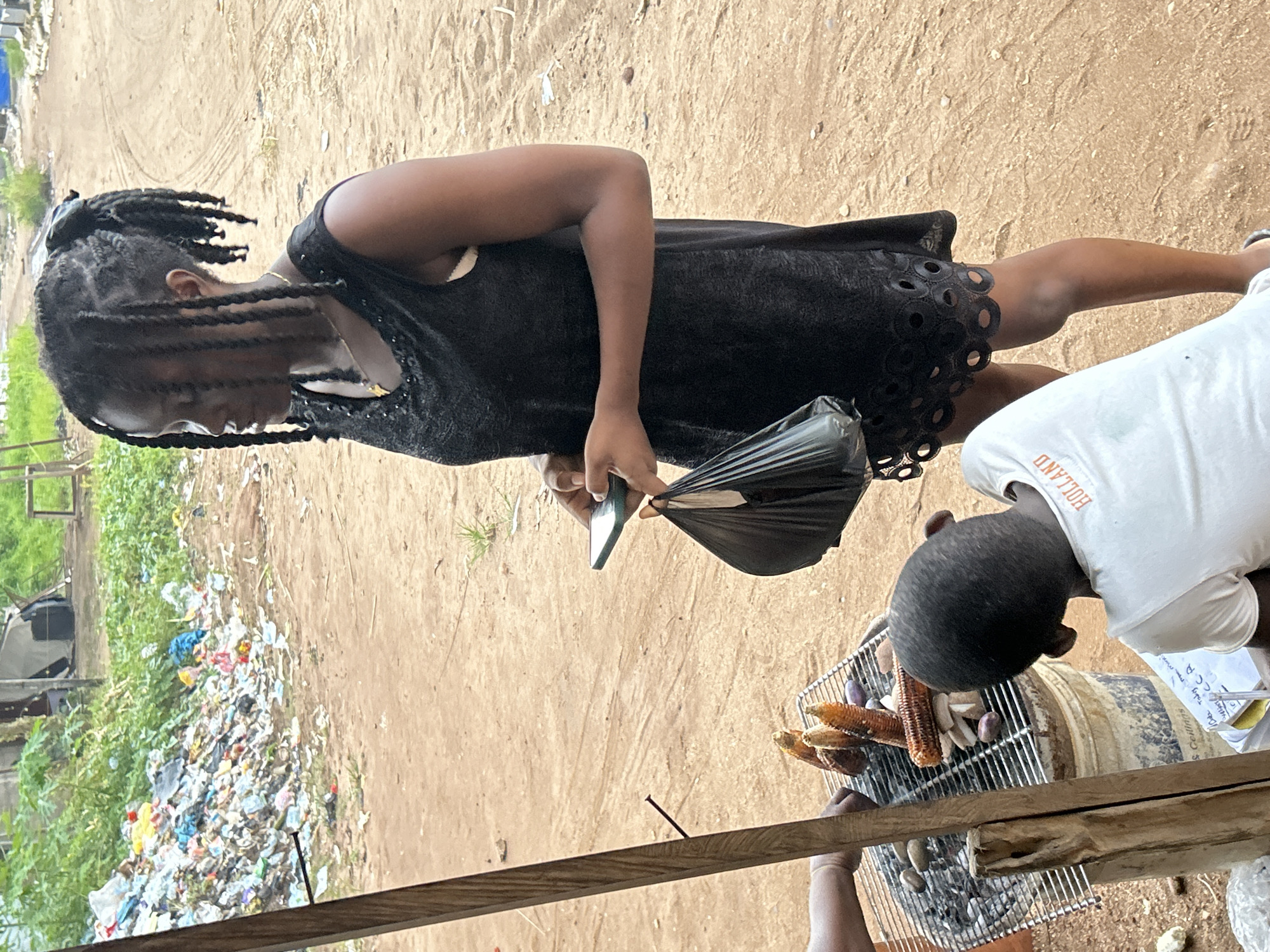 The image captures a scene outdoors, possibly in a community setting in Ihiagwa, Nigeria. Two individuals are visible, with a young child or adolescent in the foreground, wearing a white t-shirt with orange lettering that reads "HOLLAND". This child is focused on a grill situated above a bucket, where corn and other items are being cooked. A woman or older girl, with braided hair and wearing a black dress, is standing next to the grill. She is holding a black plastic bag and a mobile phone.
The setting appears to be somewhat unkempt, with a pile of trash visible in the background, mixed with greenery. The ground is sandy and dry, suggesting a warm climate. The lighting indicates it's daytime. The overall atmosphere seems casual, with the focus on the preparation of food on the grill. There's a sense of everyday life unfolding in this outdoor environment.