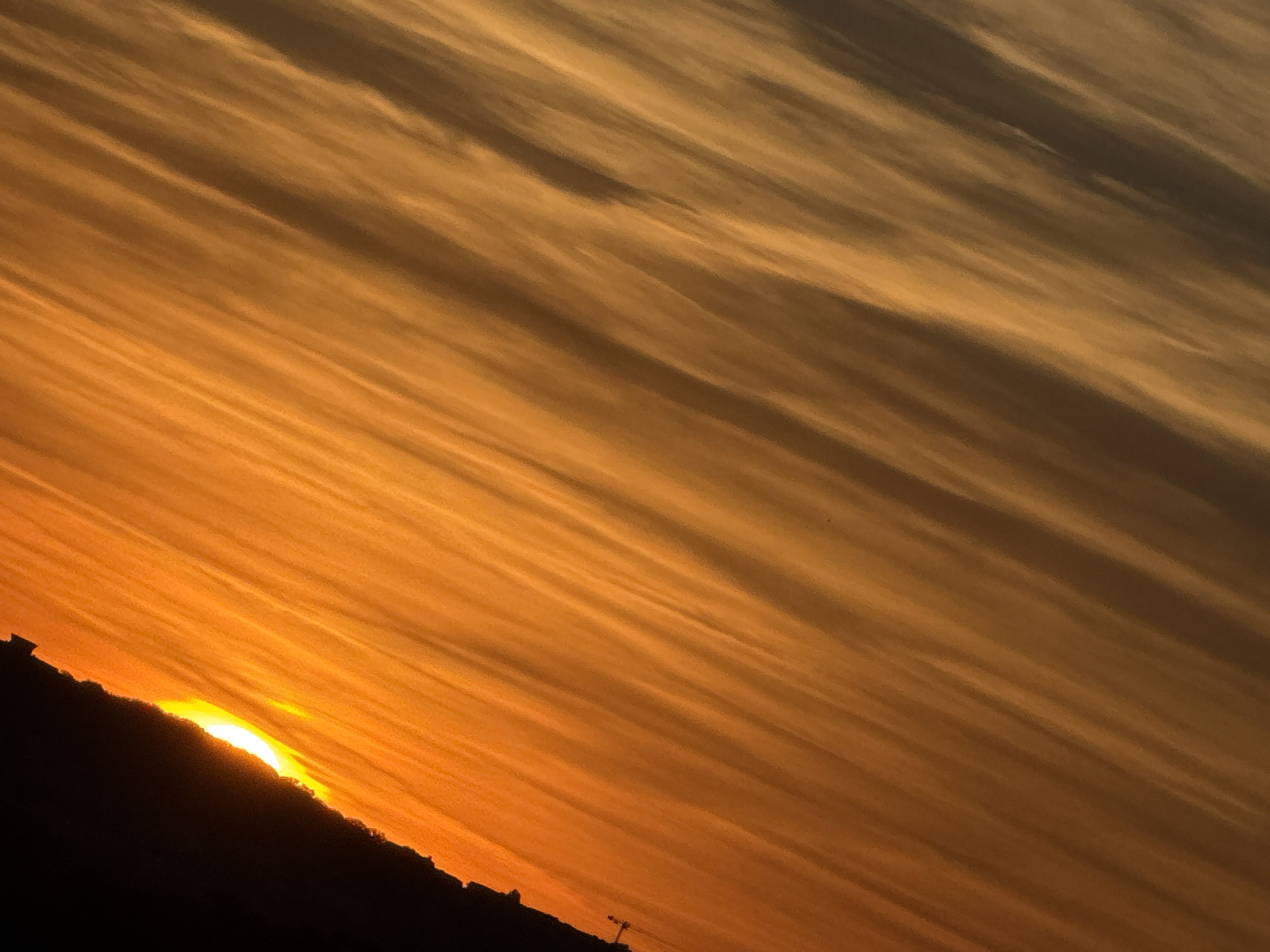 A sunset is captured from a tilted perspective, with the horizon line running diagonally from the lower left to the upper right. The sky is filled with streaks of color ranging from bright orange near the horizon to a darker, muted brown further up. A silhouetted landscape, possibly a hill or cityscape, occupies the bottom portion of the image, with a segment of the sun peeking over its edge. The sun appears as a bright, glowing orb partially obscured by the dark foreground. The overall impression is one of dramatic natural beauty.