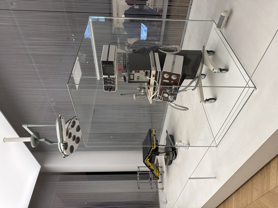 The image shows a medical display in the Wellcome Collection museum in London, located at the coordinates (51.497, -0.176). The display features a vintage medical operating table and an older model of a medical breathing apparatus. The table is made of metal and is on wheels. The breathing apparatus is enclosed in a clear glass box on a wheeled stand. The display is lit by a ceiling-mounted operating room light. The environment is sterile and white with a wooden floor. There are no people present. The objects are arranged in a symmetrical fashion and appear to be part of a museum display about the history of medicine.