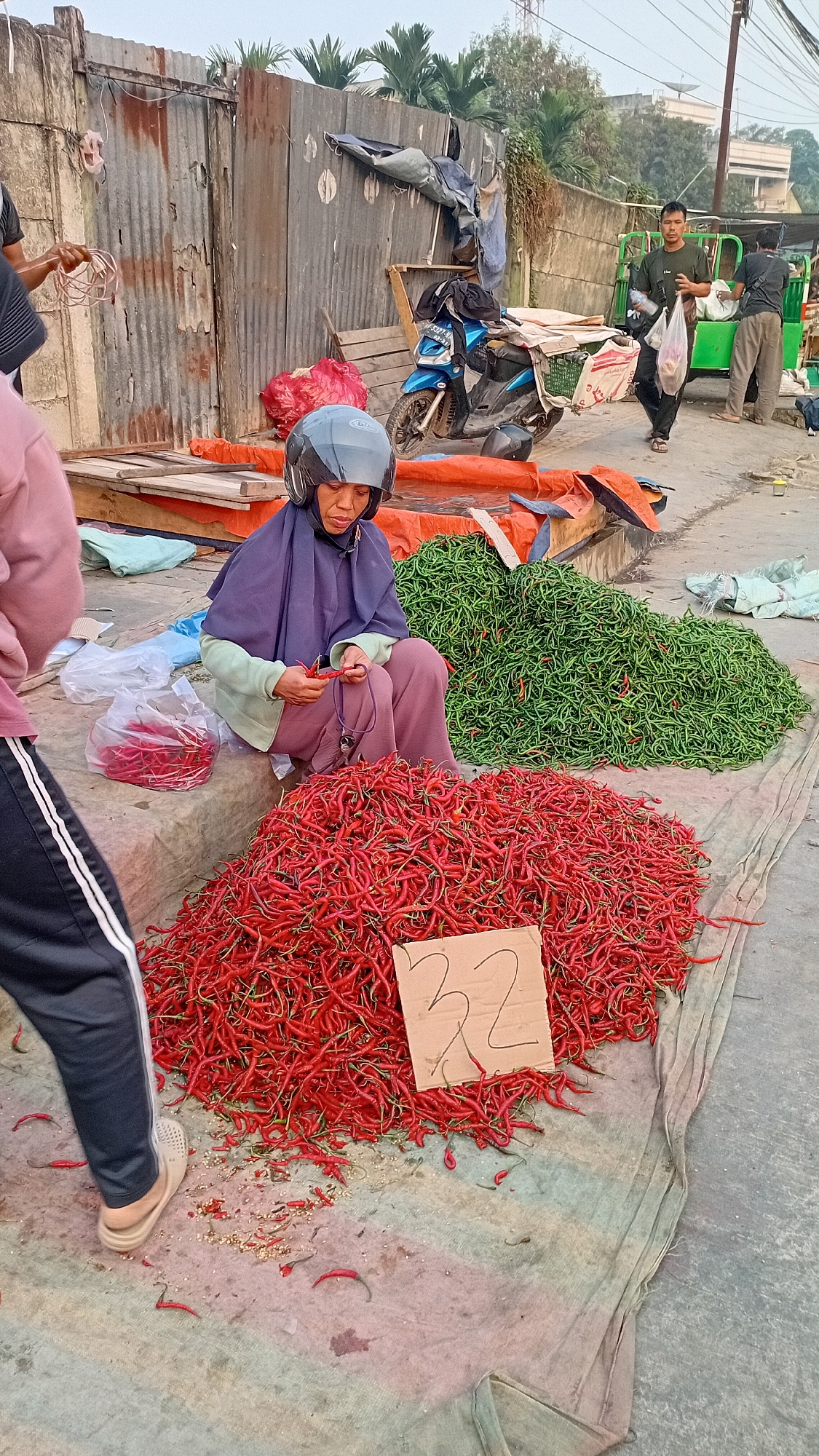 In Jambi City, Indonesia, a street market scene unfolds with a focus on fresh produce. A woman, wearing a purple hijab and a light green jacket, sits on the ground next to a large pile of red chili peppers, actively sorting through them. She is wearing a dark helmet, suggesting she arrived via motorcycle. In front of her, a cardboard sign displays the number "32". Adjacent to the red chilies is a substantial mound of green chilies. Behind the woman, a blue motorcycle is parked, and in the background, other individuals are visible, some carrying bags and interacting with what appears to be a vendor or customer, near a food stall or cart. The environment suggests a bustling outdoor market with corrugated metal walls and palm trees in the distance, indicative of a tropical or subtropical climate.