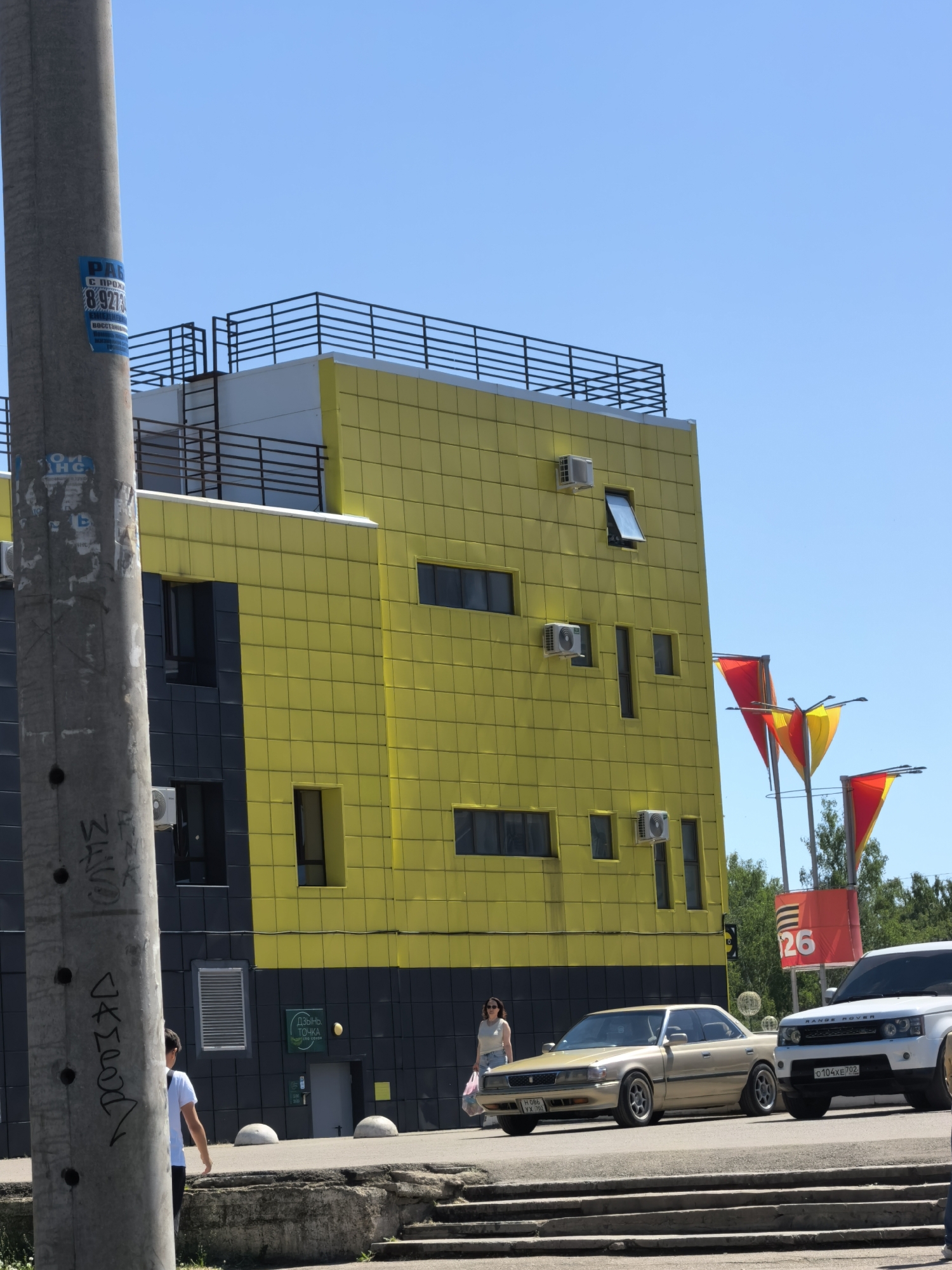 The image shows a street scene in Sterlitamak, Russia. The main focus is a yellow and black building with a modern design. Several windows and air conditioning units are visible on the building's facade. In front of the building, two cars are parked: a gold-colored vintage coupe and a white Range Rover with license plate "O 104 XE 702". A woman carrying a bag is walking on the sidewalk in front of the building, and another person is walking on the left side of the frame.
In the background, there are red and yellow flags with the number "26" on a square plate. The sky is clear and blue. A light pole covered in graffiti is visible on the left side of the frame.