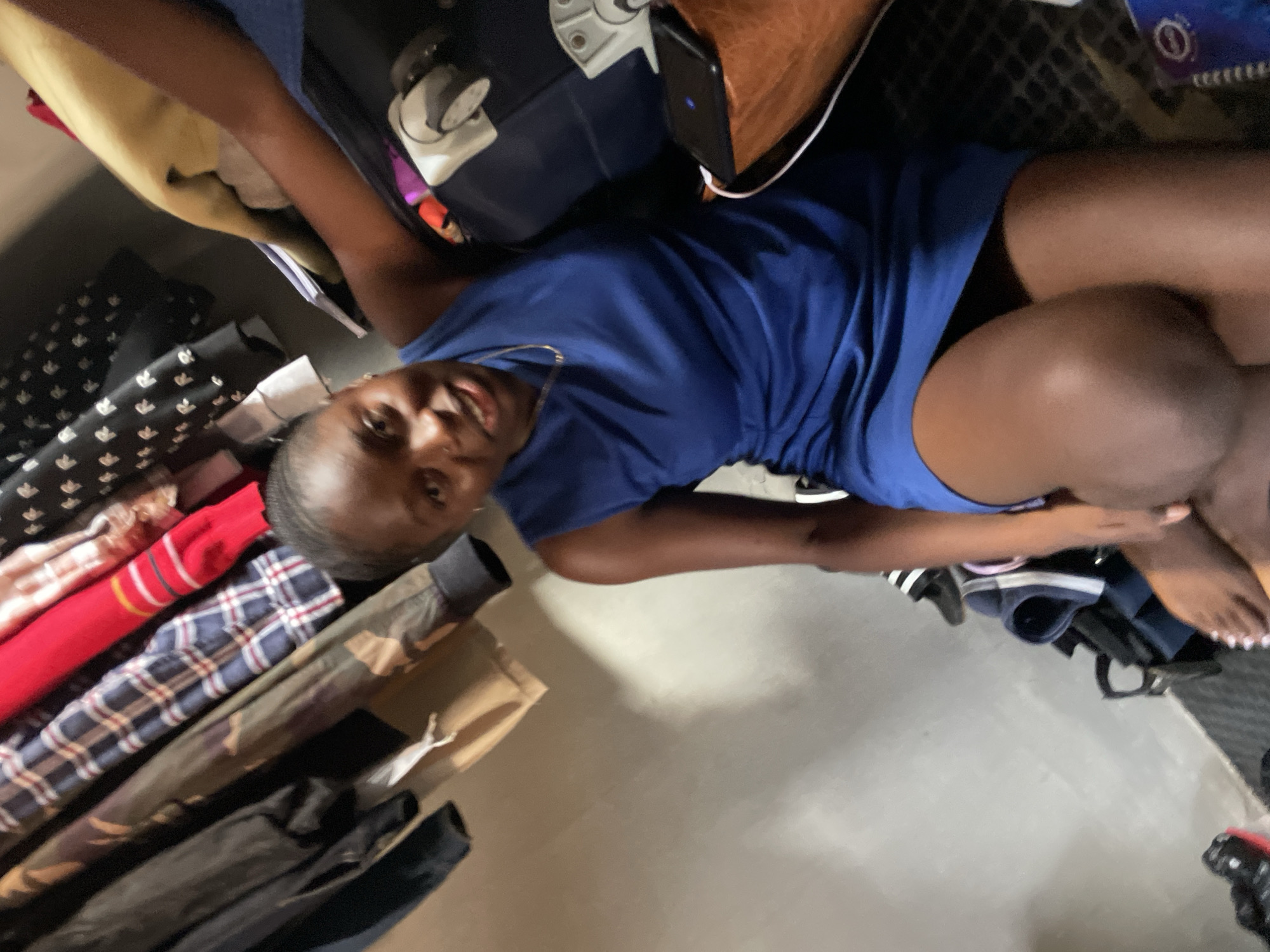 The image captures a young Black woman in a blue dress sitting on the floor of what appears to be a closet. She is looking towards the camera with a slight smile. Her legs are bent and her feet are visible, with painted toenails. Behind her, a collection of clothing is hanging on a rack, including shirts with plaid and patterned designs, as well as jackets. To her right, a brown bag and a black phone charging are visible. The environment is dimly lit and appears to be an indoor domestic setting. The overall impression is a casual and candid moment, possibly taken in Jos, Nigeria, given the context of the request.