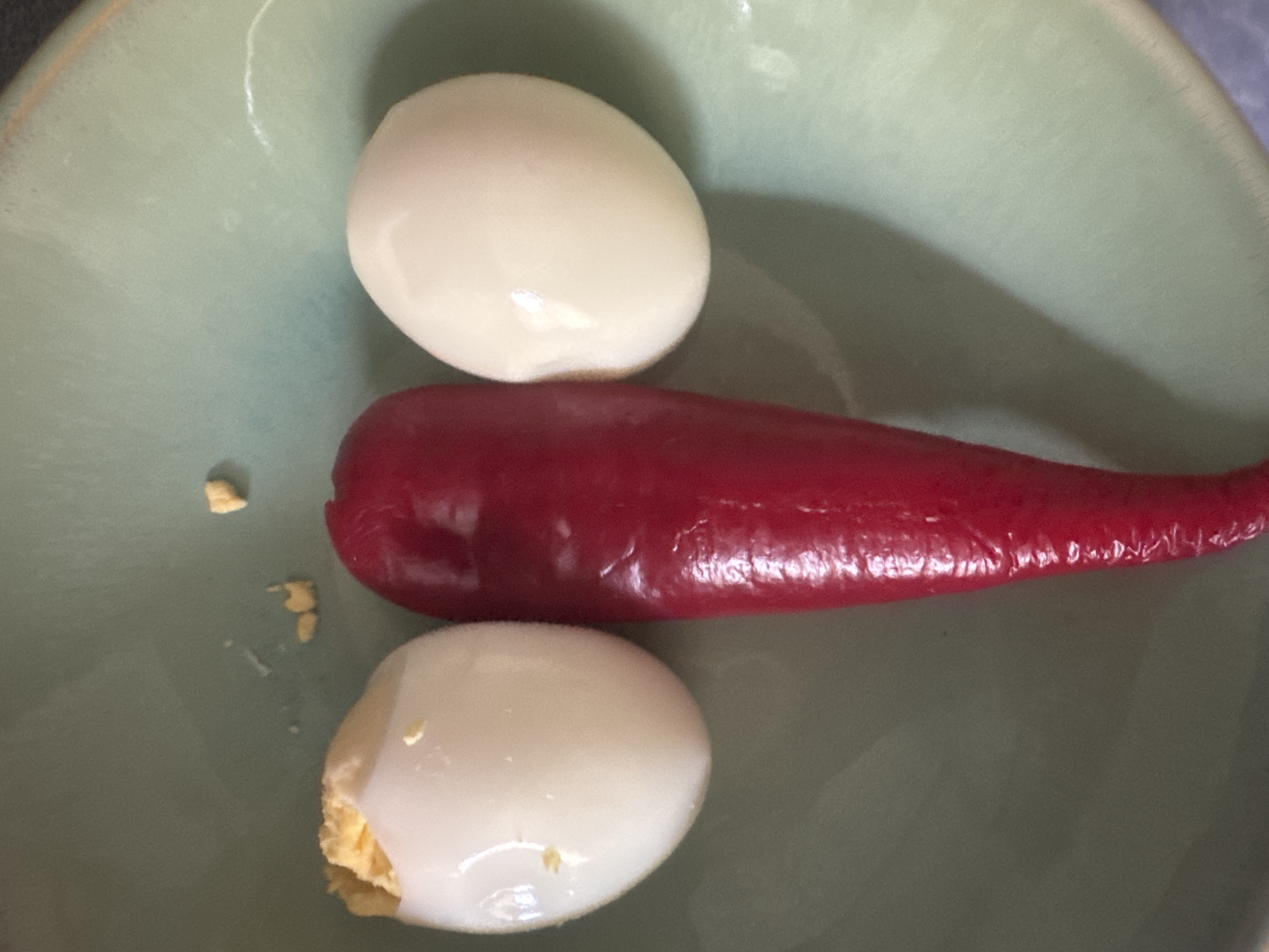 A top-down, close-up shot shows two peeled hard-boiled eggs and a red chili pepper arranged in a light green bowl. The eggs are positioned vertically, one above the other, with the chili pepper horizontally placed between them. The top egg is whole and smooth, while the bottom egg has a bite taken out of it, revealing the yellow yolk. The chili pepper is glossy and appears to be whole. Scattered on the surface of the bowl are a few small, light yellow crumbs, likely from the eggs. The lighting is soft, suggesting an indoor setting. The scene appears to be a simple preparation or presentation of food. No people, text, or specific location cues within the image itself are visible.