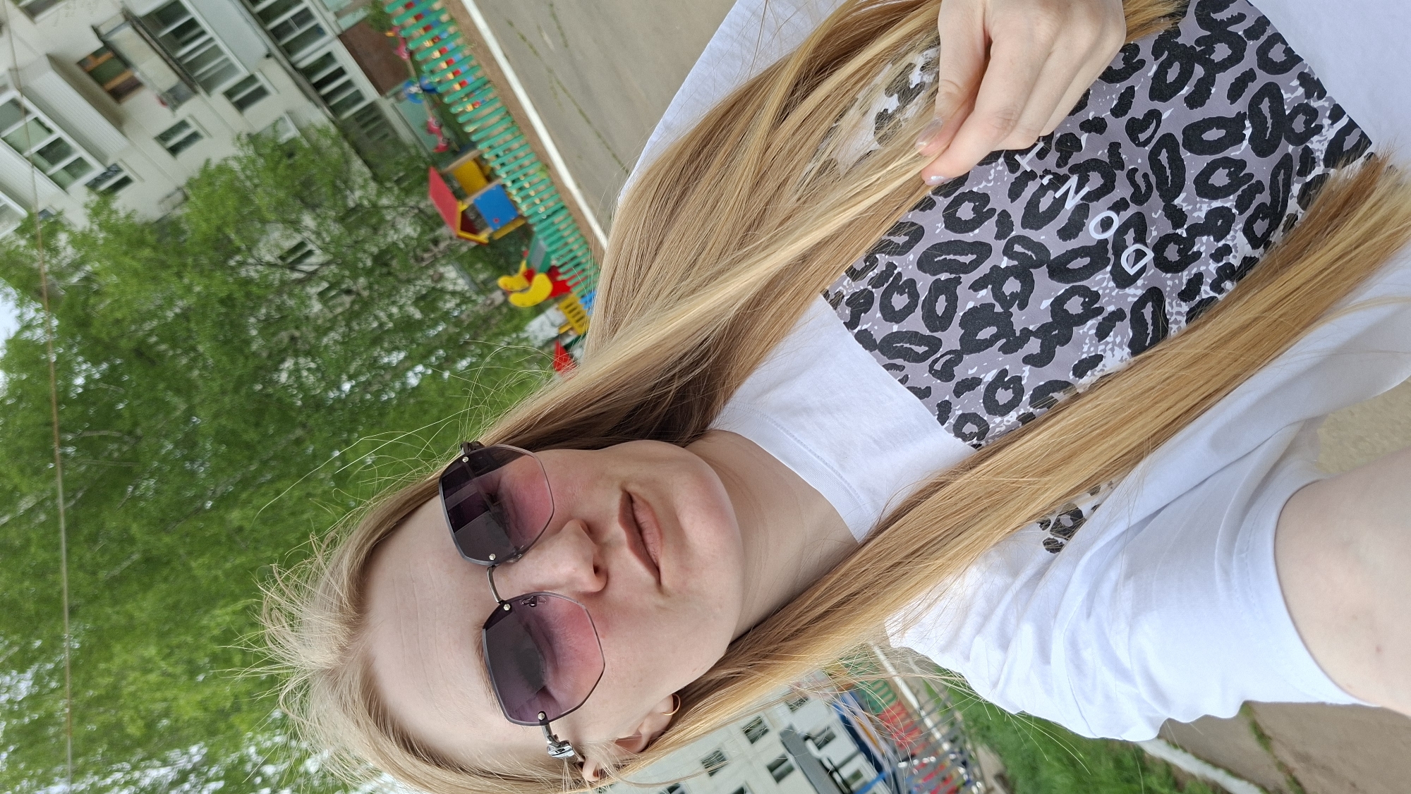 A selfie depicts a fair-skinned person with long blonde hair and sunglasses. They are wearing a white t-shirt. The person is holding a gray and black leopard print object. In the background, a playground is visible, along with apartment buildings, suggesting an urban residential area. Given the location in Chaikovsky, Russia, the setting appears to be a typical urban courtyard.