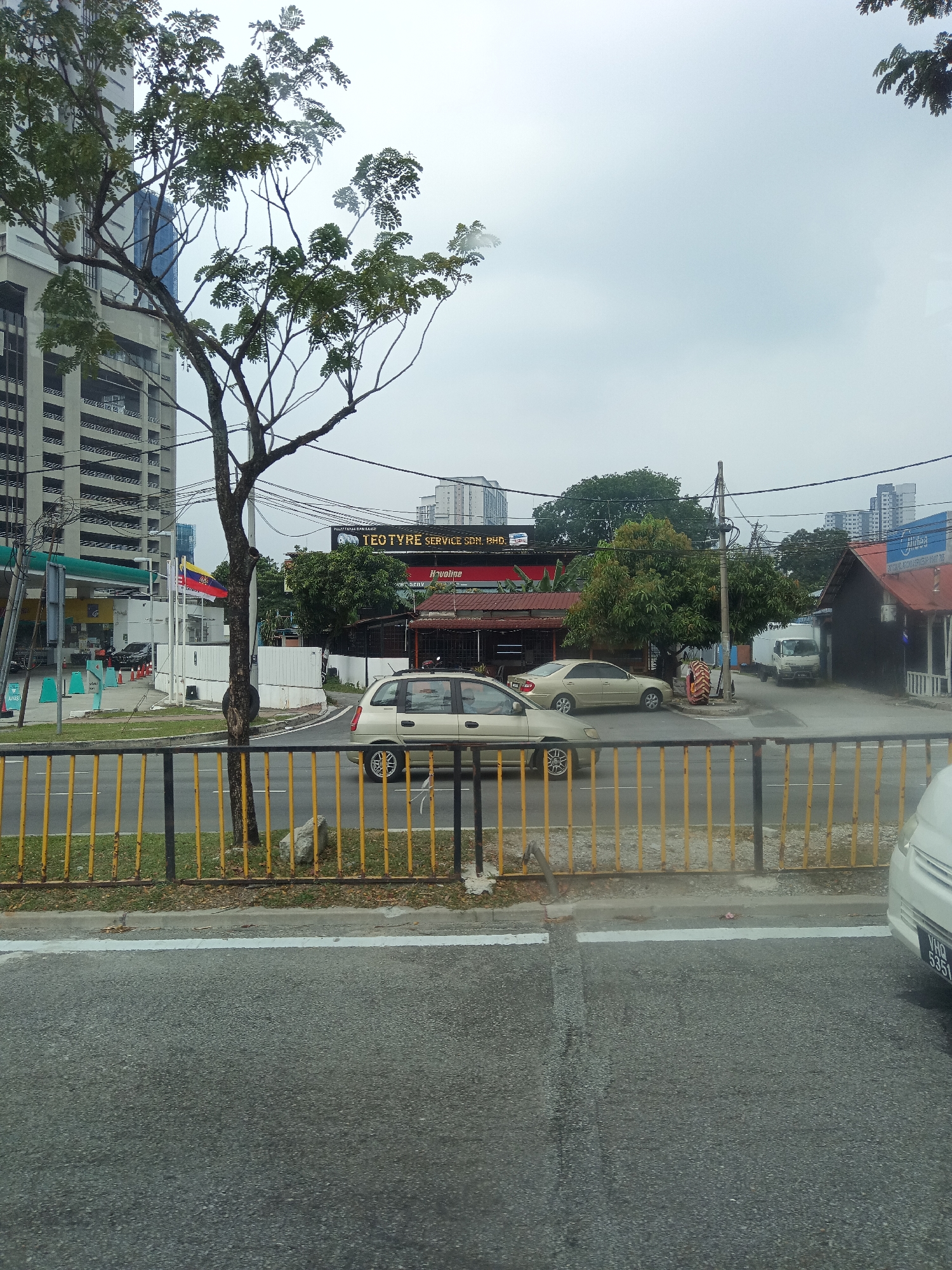 The image captures a street scene in Kuala Lumpur, Malaysia, under an overcast sky. A prominent tree with sparse leaves stands in the foreground, partially obscuring a tall, modern building with horizontal banding on the left side of the frame. A yellow and black striped fence runs along the side of the road.
On the right side of the road, a gold sedan is parked or moving slowly. Further down the street, a row of buildings with red roofs is visible, including a business with a sign that reads "TEOTYRE SERVICE SDN. BHD." Two Malaysian flags are displayed on poles near the gas station.
In the background, more buildings of varying heights can be seen, indicating an urban environment. A white delivery truck and another vehicle are also present. The road is paved asphalt with white lane markings. A white car is partially visible on the far right, its license plate reading "VHG 5351". The overall impression is that of a typical urban street on a cloudy day.