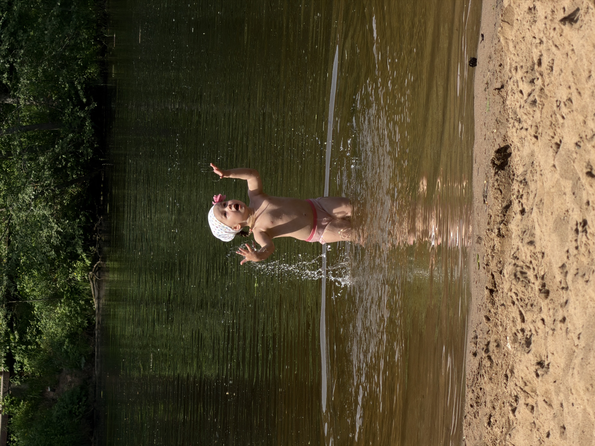 A young child stands in shallow water at the edge of a sandy beach, appearing to be in the midst of splashing or playing. The child is wearing a light-colored bonnet with a pink flower detail, and pink bottoms. The water line is just above the child's knees, creating splashing effects around the lower body. The background includes lush green vegetation on one side and a sandy beach on the other. It is not possible to determine the specific location in Moscow, Russia, based on the image alone.