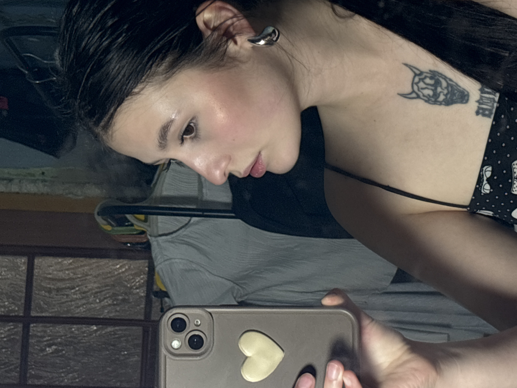 A woman is taking a selfie in an indoor environment. She has dark hair that appears to be wet or styled with product. Her skin is fair, and she is wearing a silver earring on her left ear. There is a tattoo of a demonic head on her chest, along with some script. She is holding a mobile phone in her right hand, and the phone has a light-colored heart-shaped sticker on its back. Behind her, there is a reflection of a window with a grid pattern, and a glimpse of some clothing or textiles. It is impossible to determine the exact location of the scene in the city of Фоки, Russia from the image alone.