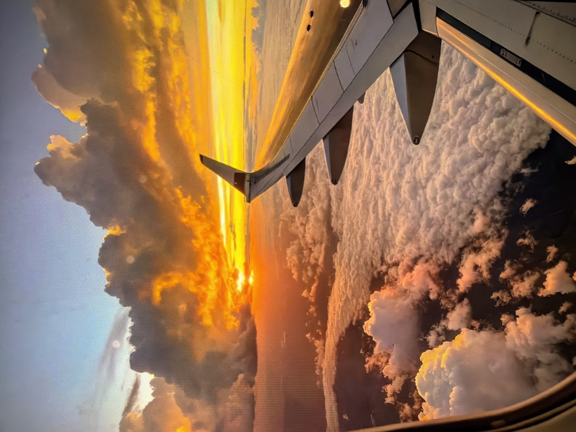A view from an airplane window shows the wing extending over a thick cloud cover, with a sunset visible in the distance. The wing of the plane is white, with visible flaps. The clouds are dense and extend into the horizon. The sunset is a gradient of yellow, orange, and red, casting a glow over the sky and clouds. The location and city are not determinable from the image.