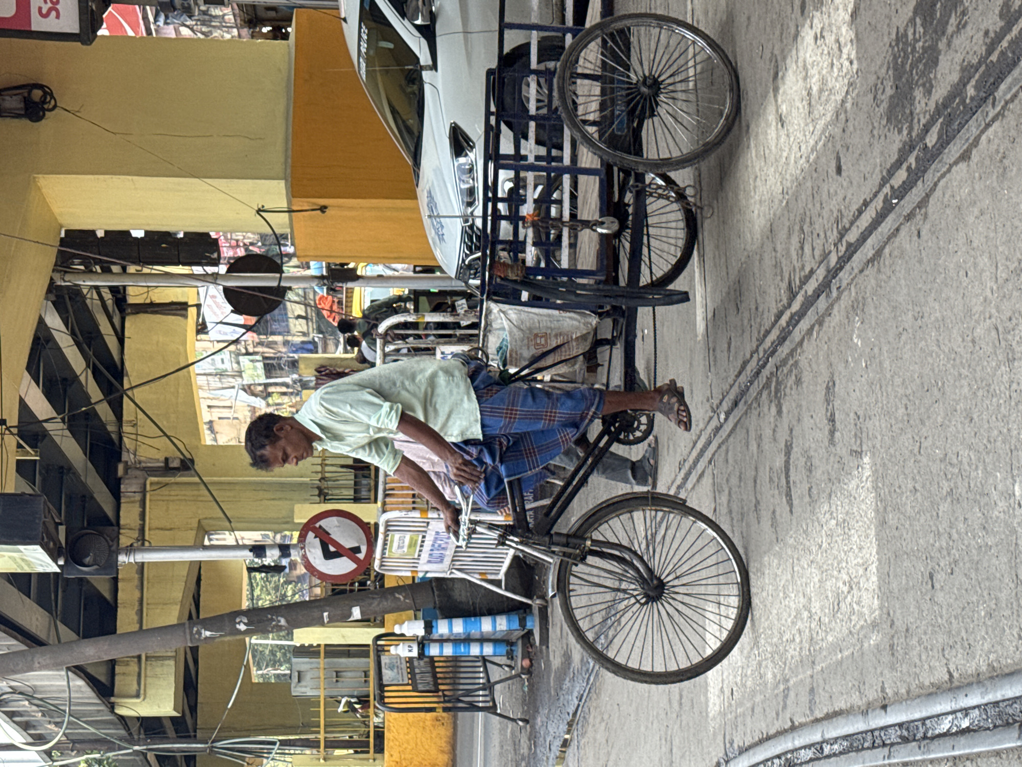 In Kolkata, India, a man pedals a rickshaw laden with a white vehicle atop its frame. The man wears a light green shirt and blue plaid pants, with sandals on his feet. A “No Left Turn” sign is visible behind the rickshaw. The setting appears to be a street with buildings and infrastructure nearby.