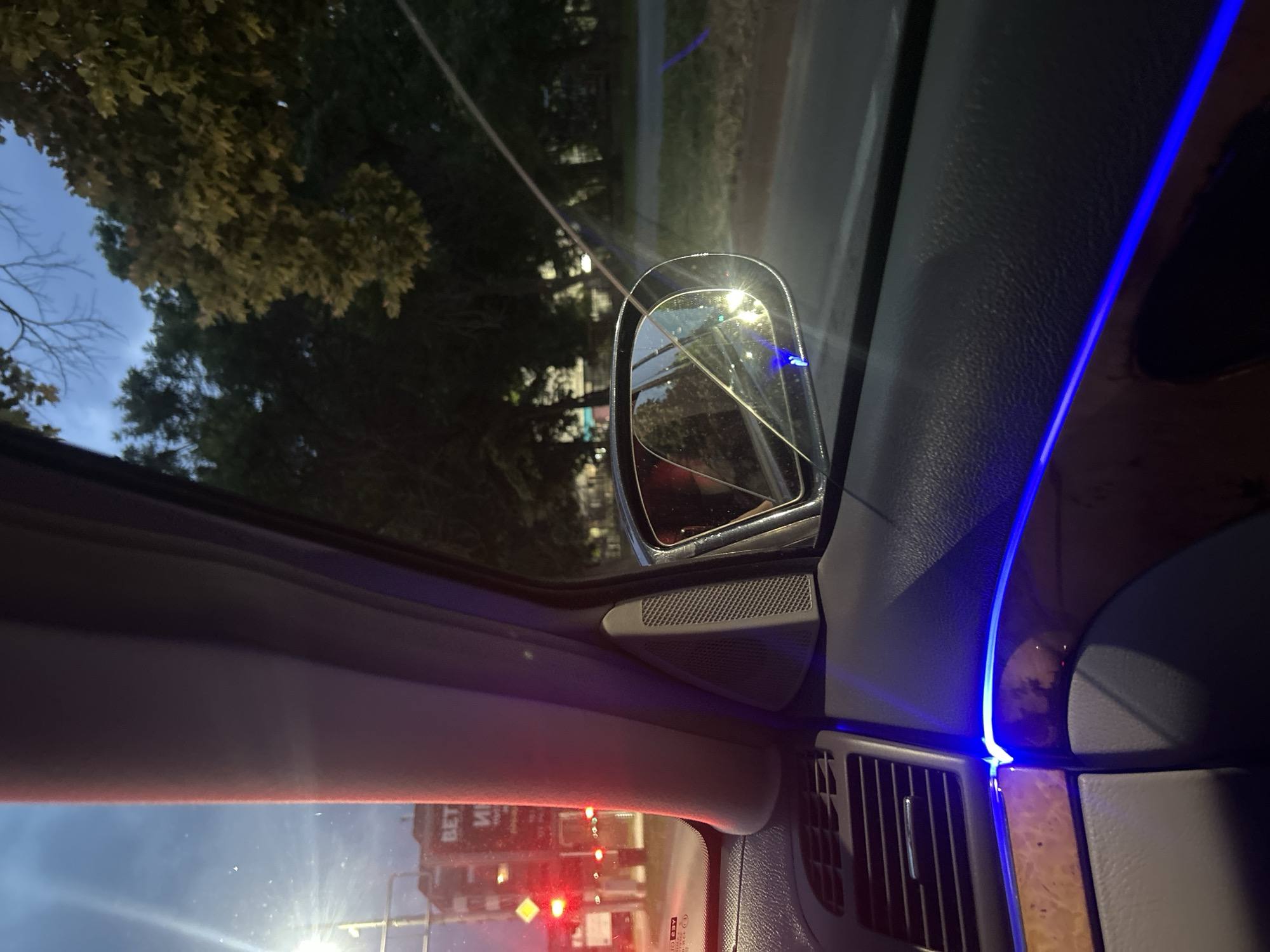 The image captures the interior of a passenger vehicle at dusk or night, viewed towards the front passenger side. Blue ambient LED lighting runs horizontally across the dark gray dashboard and a segment of wood trim. An air vent is visible on the left of the dashboard. The vehicle's A-pillar is prominent in the left-center, appearing light gray. A speaker grille is integrated into the passenger side door panel near the A-pillar.
Through the front passenger window, an urban environment in Plovdiv, Bulgaria, is visible under a dark blue sky. Multiple red traffic lights are illuminated on the left, indicating the vehicle is stationary at an intersection. In the background on the left, a building displays illuminated Cyrillic text, clearly showing "ВЕТ И".
The exterior rearview mirror on the passenger side is visible, exhibiting a distinct diagonal crack or line across its reflective surface. The mirror reflects interior lighting and a blurred view of the road behind. Beyond the mirror, a paved road extends into the distance, lined with dark green deciduous trees. A faint blue glow, originating from the vehicle's interior lighting, is reflected on the ground outside, possibly indicating a road marking or path. No individuals are identifiable within the frame. The scene is illuminated by artificial light sources contrasting with the ambient darkness.