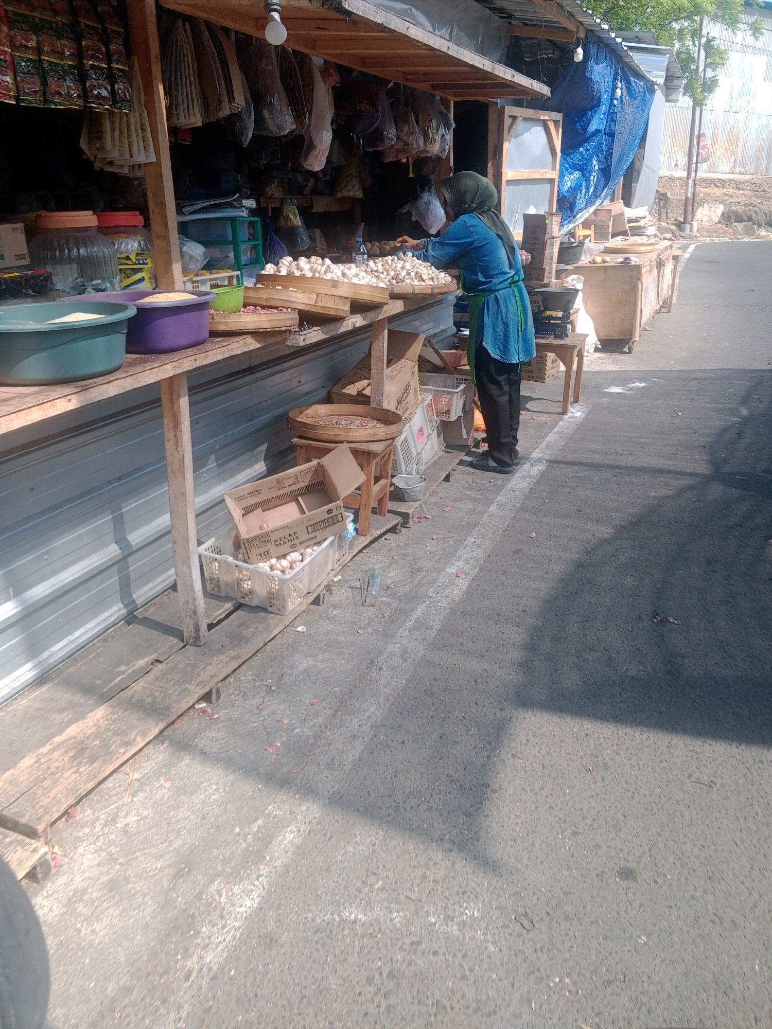 A woman wearing a blue headscarf and a blue tunic over black pants is standing at a roadside stall in Wonogiri, Indonesia. She appears to be organizing or displaying garlic bulbs from a woven tray. The stall is set up under a wooden awning covered with a blue tarp. Various goods, including packets of spices, bags of produce, and containers of rice, are visible on shelves and in baskets. To the right of the stall, a road with a white line marker is visible, with some scattered debris on the ground. The overall scene depicts a typical market setting in a semi-urban or rural area.