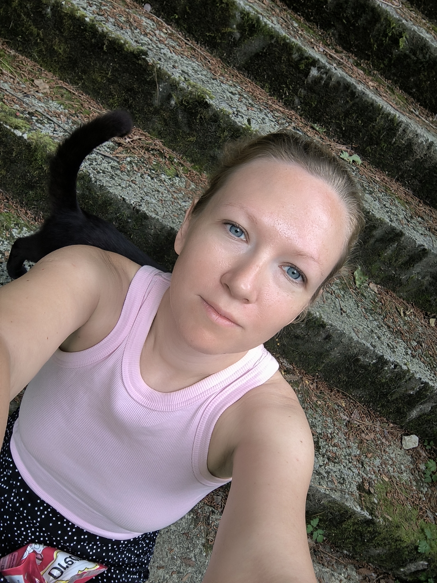 A woman with fair skin and blonde hair, wearing a pink ribbed tank top and a black and white polka dot skirt, is taking a selfie on an outdoor stone staircase covered in moss and fallen leaves. A black cat is partially visible behind her. The staircase appears to be in a natural, possibly wooded, setting.
