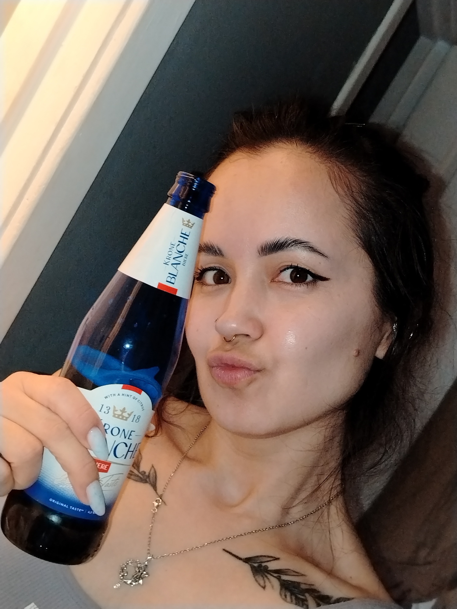 An indoor, close-up shot features a young woman holding a blue glass bottle, likely a beer, to her right cheek. She is looking directly at the camera with a playful expression, her lips puckered into a "kissy face." Her dark hair is pulled back, and she has visible eyeliner, a nose ring (gold-colored hoop), and small earrings. Her long nails are painted a light, possibly pale blue or grey color. A delicate silver necklace with a decorative pendant is visible around her neck, and a tattoo resembling a leafy branch is on her upper chest/shoulder area.
The bottle she holds is labeled "KRONE BLANCHE BIERE" with a crown logo. Additional text on the label reads "WITH A HINT OF CITRUS," "13 18," and "ORIGINAL TASTE." The background is softly lit, showing vertical striped patterns on the left, possibly from blinds or wall paneling, and a darker, indistinct area to the right, suggesting an interior setting, possibly a home in Noviy, Russia. The lighting appears soft, indicating evening or nighttime.