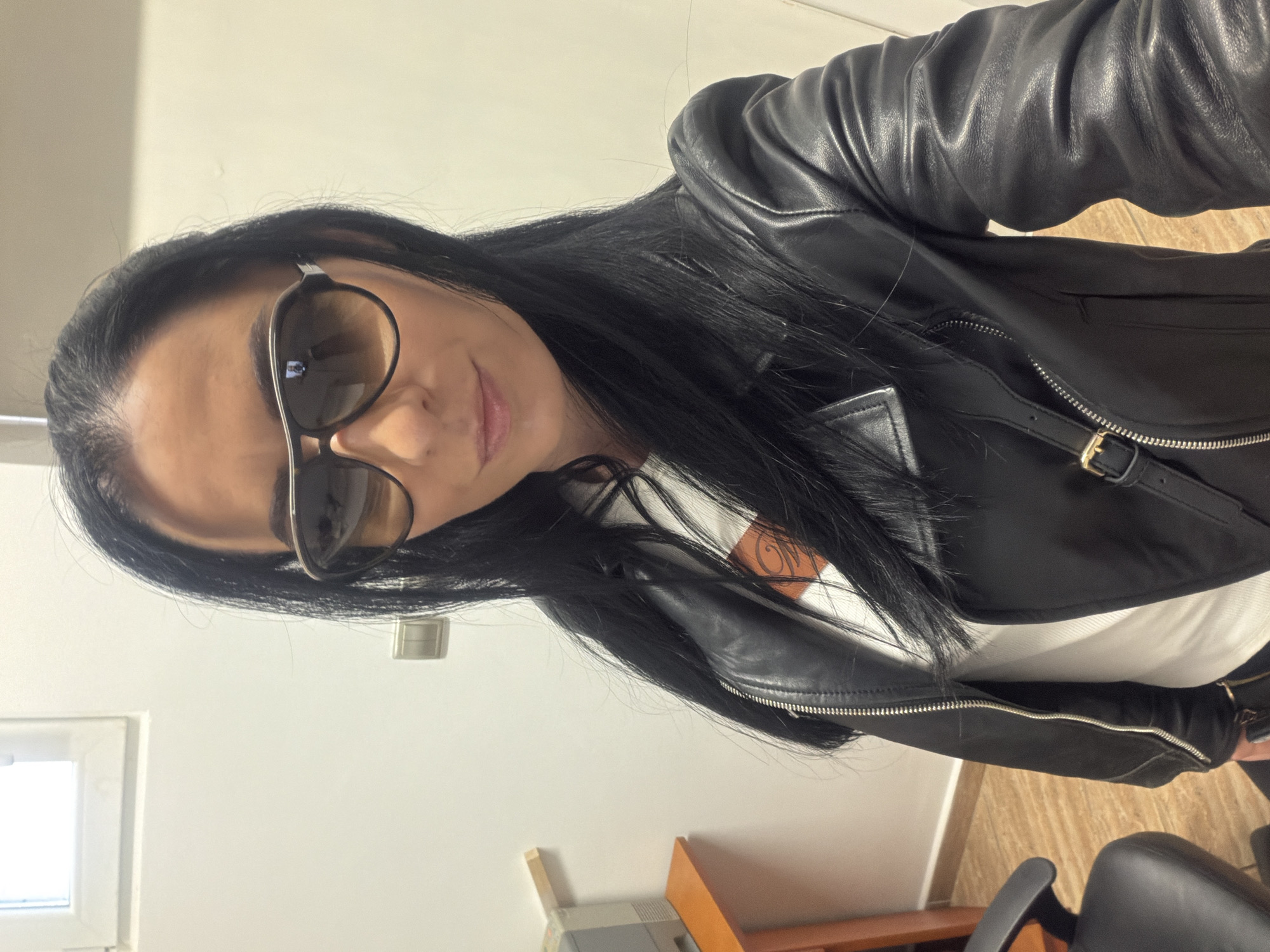 The image captures a female individual, likely in her 30s or 40s, posing for a self-portrait indoors. She has long, dark, straight hair and wears dark aviator-style sunglasses with gradient lenses. Her lips are slightly parted in a faint smile. She is dressed in a black leather motorcycle-style jacket, partially unzipped, revealing a white top underneath with a subtle reddish-brown graphic detail on the chest. A small gold-colored buckle is visible on a strap near the right shoulder of the jacket.
The scene takes place within an interior room, suggestive of an office or private workspace in Pleven, Bulgaria. The background consists of plain, light-colored walls. In the upper left corner, a white-framed window is visible, providing a source of natural light. Below the window and to the left, a section of an orange or light brown wooden desk or shelf is present, topped with a grey office printer or similar equipment. A light-colored double switch or electrical outlet is mounted on the wall behind the individual's right shoulder. In the lower left quadrant, a portion of a black office chair and a light brown floor, possibly wood or laminate, are visible. No specific architectural landmarks or signs are present to precisely identify the building or street within Pleven.
