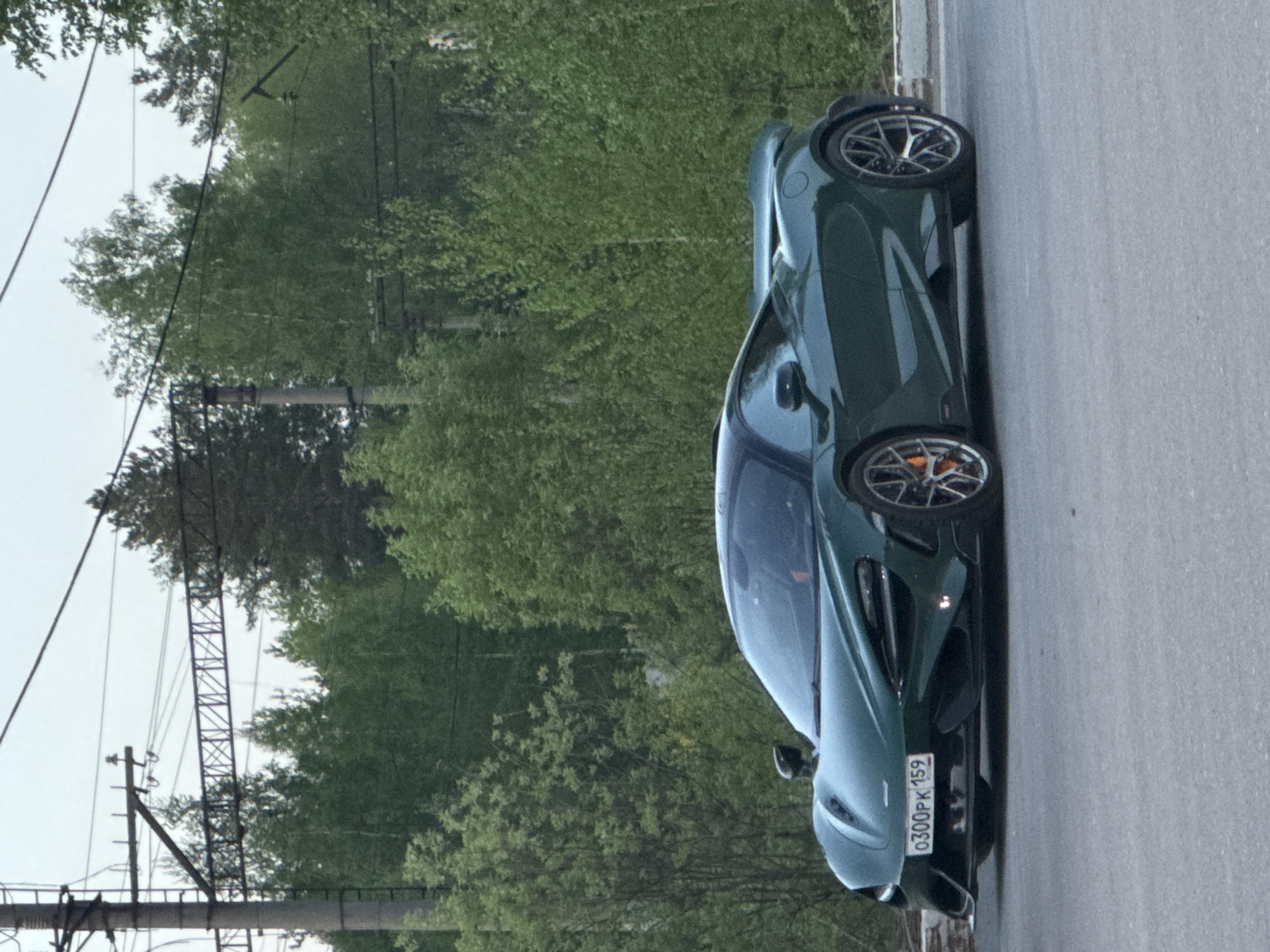 The image features a green McLaren sports car parked on the side of a gray asphalt road. The license plate is visible and reads "0300PK 159," which indicates the car is registered in the Perm Krai region of Russia. The car is facing towards the bottom of the image. In the background, there is lush green foliage suggesting the location is likely a suburban or park area. An electrical transmission tower is visible in the background. The overall lighting suggests an overcast day.