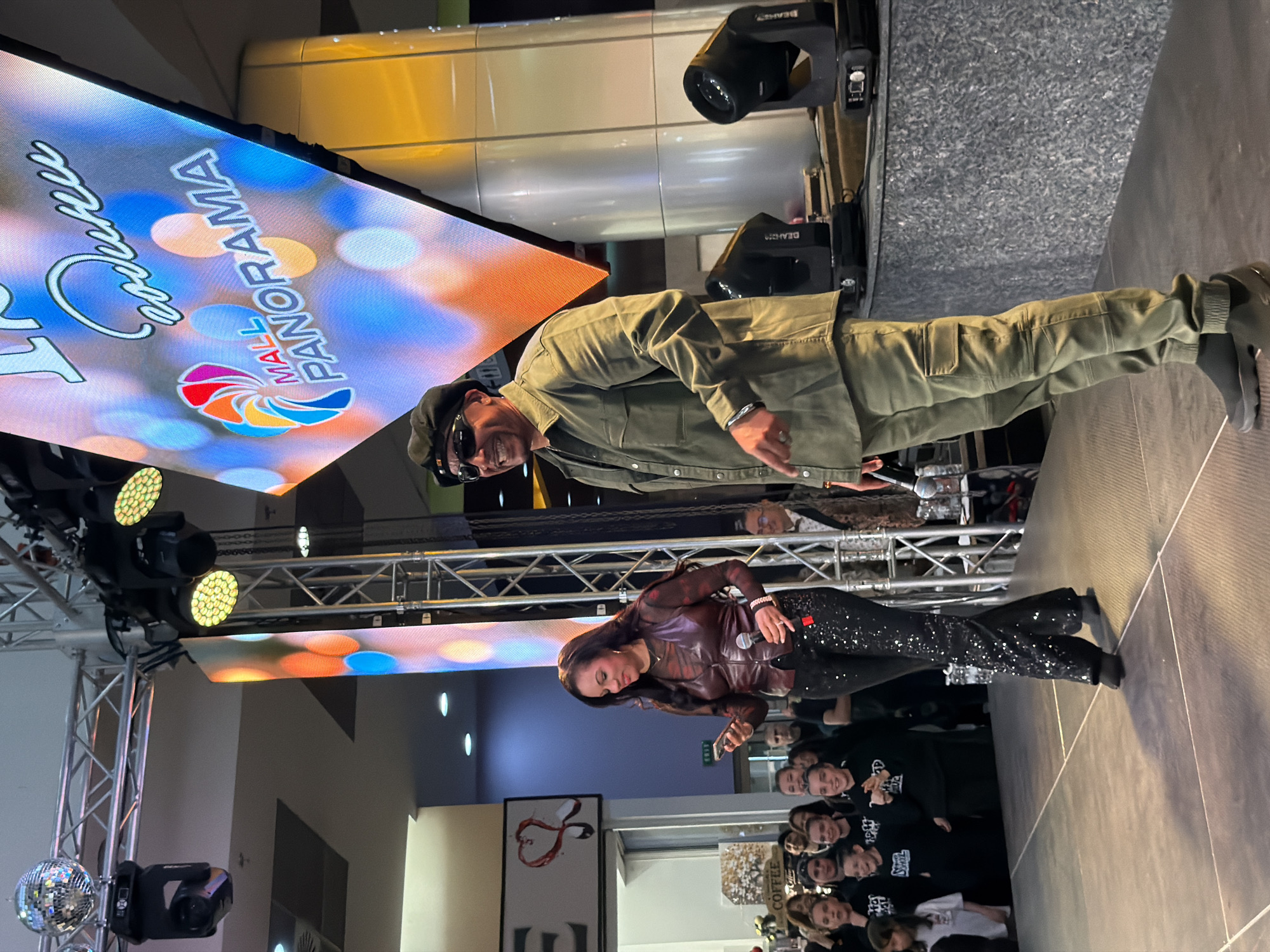 This image depicts an indoor public event, likely a performance or presentation, taking place on a stage at "Mall Panorama" in Pleven, Bulgaria.
Two main individuals are on the stage. On the right, a man with a smiling face is dressed in a khaki-colored utility jacket, matching cargo pants, dark sunglasses, and a black cap. He is seen gesturing with his right hand, which sports a metallic bracelet and a ring. To his left, a woman holds a microphone in one hand and a smartphone in the other. She is wearing a dark red/burgundy leather-like jacket over a sheer patterned top, paired with sparkling black trousers, and has long, dark hair.
The stage is professionally set up with metal trussing supporting various stage lights, including LED panel lights and moving head lights branded "BEAM7." A disco ball also hangs from the truss structure. A large LED screen serves as a dynamic backdrop, prominently displaying the text "MALL PANORAMA" with a colorful, abstract logo. Another cursive word, possibly "Cremall" or "Creamall," is also visible on the screen.
Below the stage, a cheerful audience is partially visible, with many smiling faces looking up at the stage. Some audience members are wearing dark t-shirts. In the background, hints of the mall environment are present, including a sign with "COFFEE" clearly visible and another sign featuring a red heart logo. The atmosphere appears lively and engaging, typical of a public event or show.
