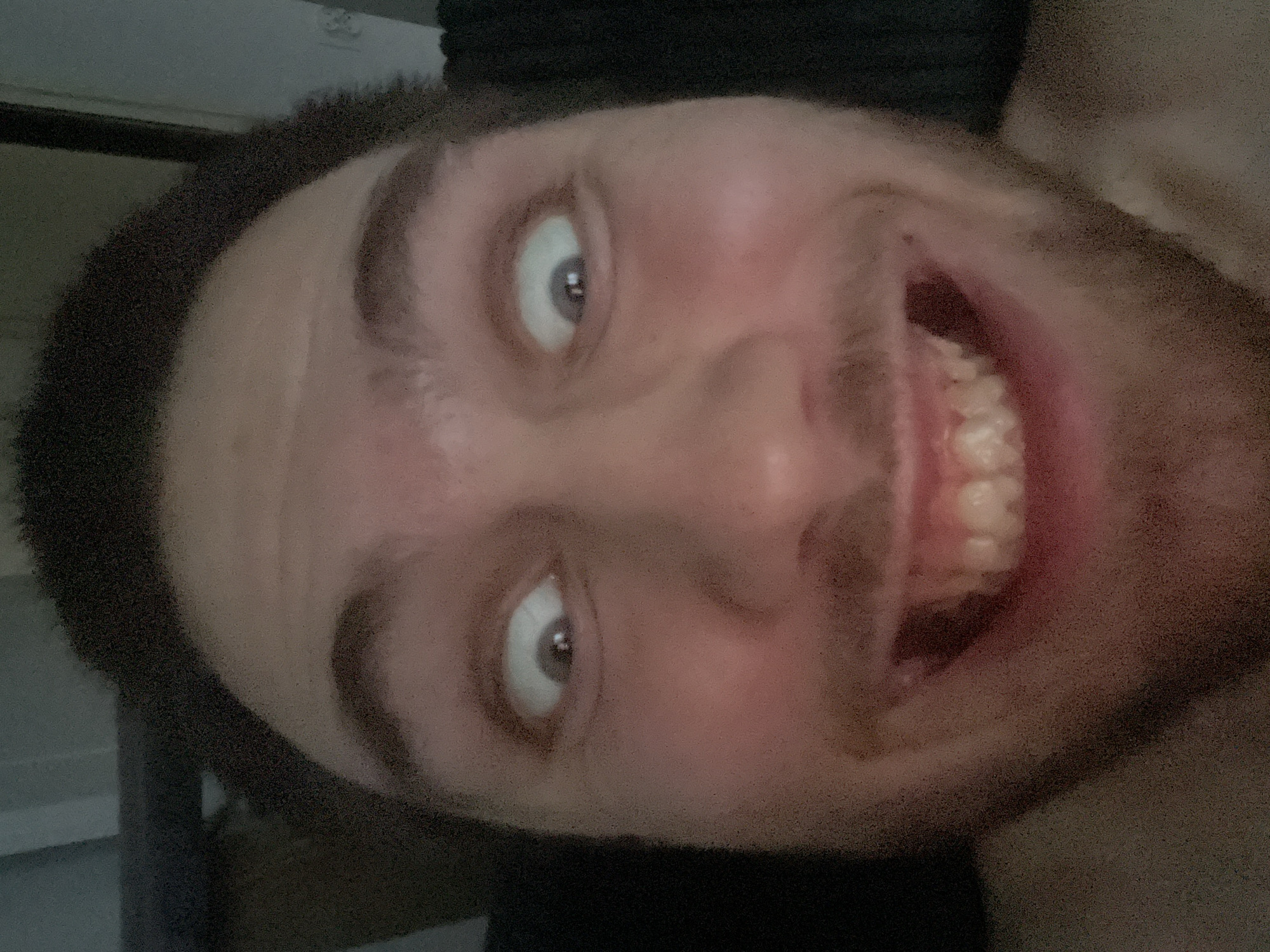 This is a close-up, rotated photograph of a person's face. The subject is looking upwards and towards the camera with a wide, open-mouthed smile, revealing their teeth. Their eyes are open, and their gaze is directed slightly upwards. The lighting appears dim, suggesting it may be nighttime or an indoor setting with low light. The background is dark and out of focus, offering no clear details about the setting. The image is grainy, indicating a possible low-resolution camera or poor lighting conditions. The orientation of the photo is such that the subject's head appears to be tilted to the right. No text is visible in the image.