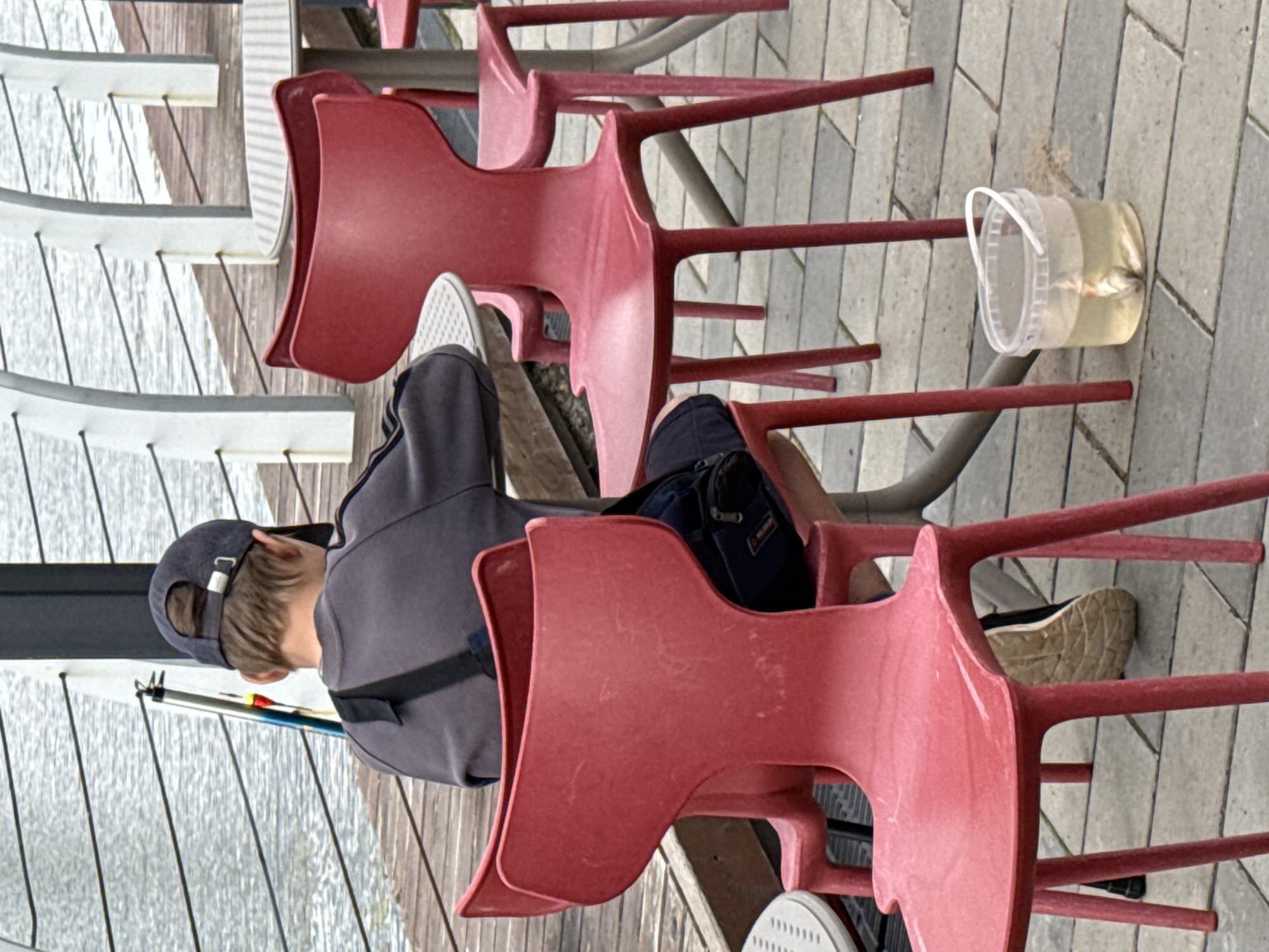 The image, rotated correctly, shows an outdoor scene featuring a young person seated at a table, seemingly engaged in fishing or having recently fished.
**Subjects:** The primary subject is a person, seen from behind, wearing a dark blue baseball cap, a grey long-sleeved top (possibly a sweatshirt or light jacket), and dark trousers. A dark blue shoulder bag is visible resting on their lap or next to them. Their feet, wearing white sneakers with grey soles, are visible under the table. Several red, lightweight plastic chairs are prominent; some are arranged around a small, dark-colored table, while others appear stacked or unused nearby. On the paved ground, next to one of the chair legs, is a small, clear plastic bucket with a white handle, containing water and at least one small fish. A fishing rod, with colorful floats or lures near its tip, is propped up next to the person, leaning against the railing.
**Scene and Setting:** The setting appears to be an outdoor paved area, possibly a promenade or pier, adjacent to a body of water (likely a lake or river, consistent with Votkinsk, Russia). A white metal railing separates the paved area from the water. The ground is covered with grey pavers laid in a geometric pattern. Wooden planks are also visible, suggesting a walkway or part of the structure over the water.
**Activity:** The person is sitting, likely fishing or observing the water, with the fishing rod nearby and the catch in the bucket. The arrangement of chairs suggests a casual outdoor seating area where this activity is taking place.
**Notable Details:** The light is diffused, suggesting an overcast day or indirect sunlight. The red chairs provide a strong color contrast against the grey pavement and white railings. The small fish in the bucket indicates a recent successful catch. No visible text is discernible in the image.
