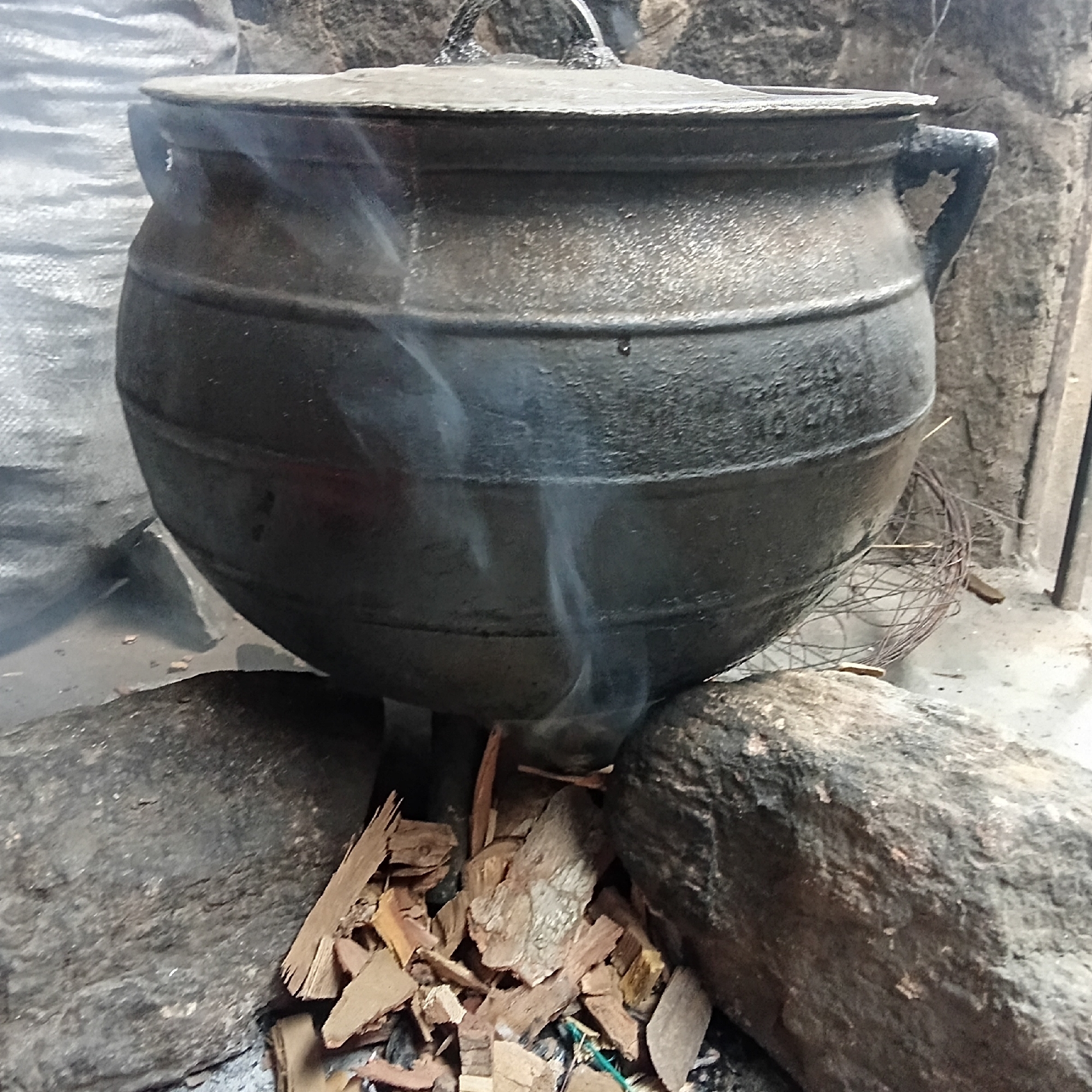The image shows a large, dark, lidded cast iron pot sitting over a fire built between three rough, dark rocks. The pot is spherical with raised horizontal bands around its middle and a handle on one side. A lid covers the top of the pot, and smoke rises from around the pot, suggesting the fire is lit. The fire consists of small pieces of wood and possibly other combustible materials. The rocks are irregular in shape and appear weathered. The background is somewhat indistinct, with a woven or mesh object and what may be a wall or other structure partially visible. The scene appears to be an outdoor cooking setup in a rustic or undeveloped location, but the city and specific region are indistinguishable.