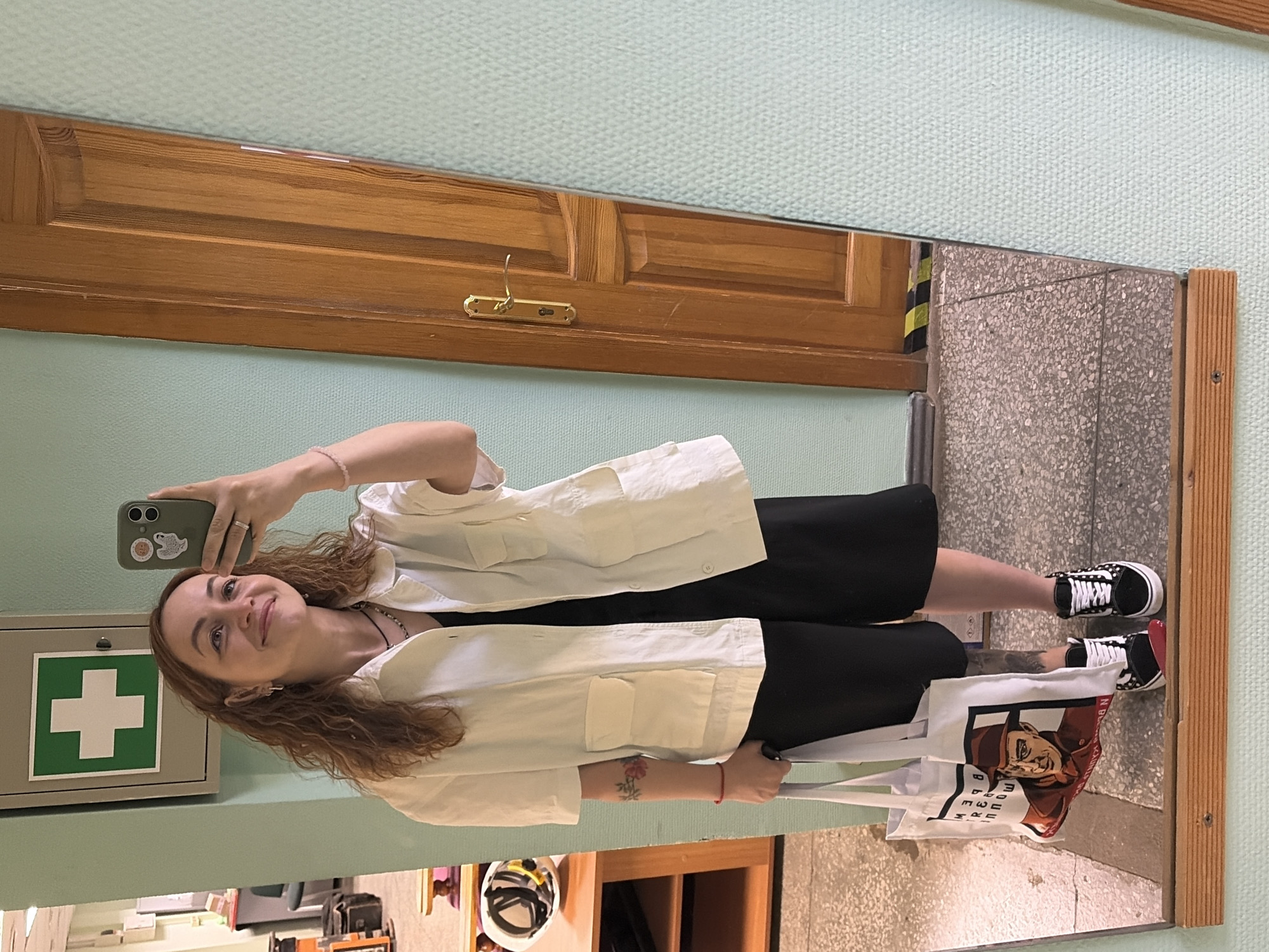 A woman is standing in front of a mirror, taking a selfie with her smartphone. She is wearing a white button-down shirt, a black dress, and black and white sneakers. She is holding a white tote bag with a graphic print on it. The mirror reflects a hallway with light blue walls, a wooden door, a first aid kit box, and a tiled floor. Based on the information available, the location is the city of Chaikovsky, Russia.