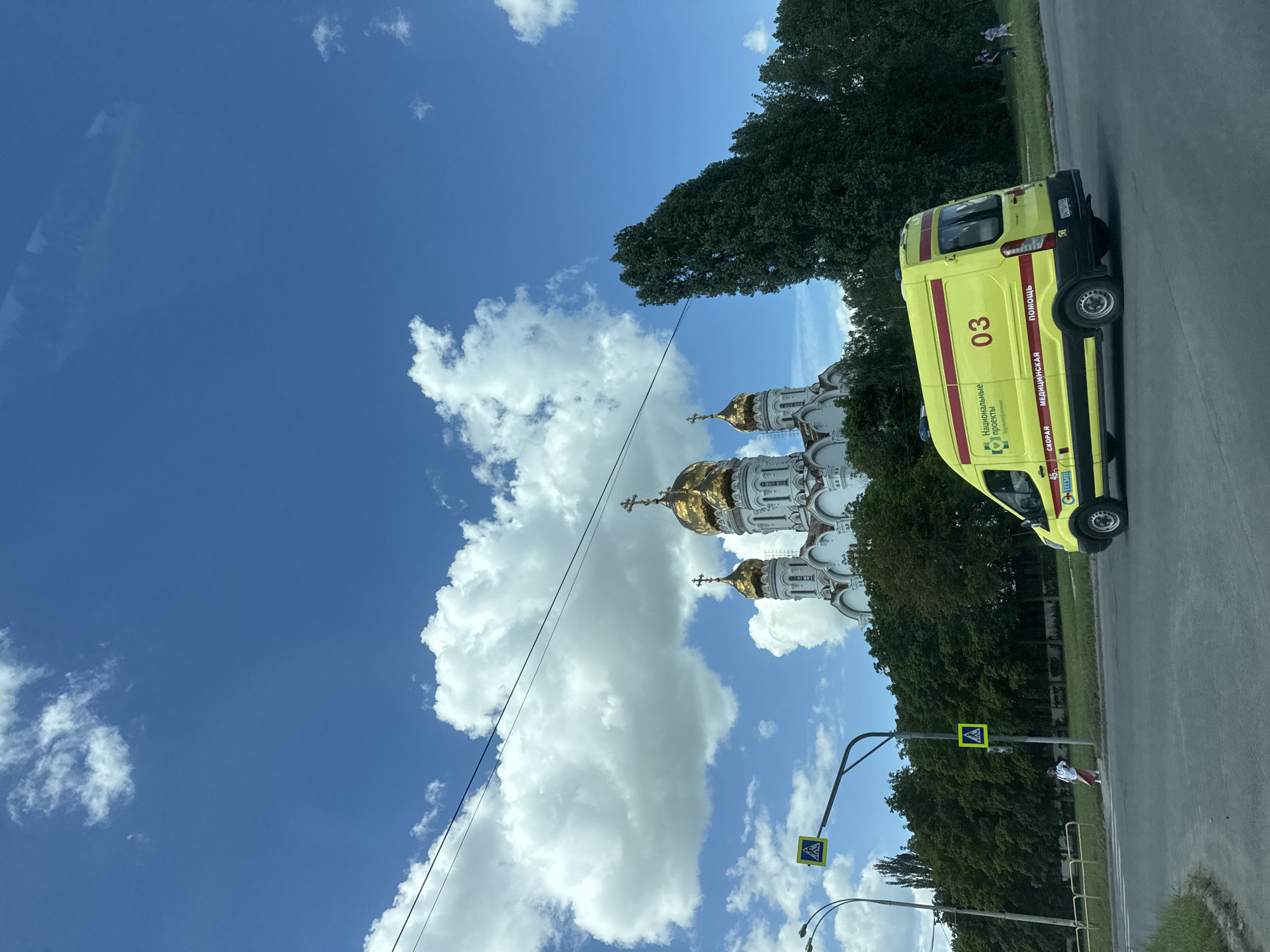 This image captures a bright, sunny day with a clear blue sky dotted with fluffy white clouds. In the foreground, a yellow ambulance with red stripes is parked on the side of a paved road. The ambulance has the number "03" prominently displayed and text that appears to be in Cyrillic script.
To the left of the ambulance, a large, white building with ornate architecture and golden domes, likely a church or cathedral, is visible. Lush green trees line the area between the road and the building, and beyond that, more trees and greenery can be seen. A pedestrian crossing sign with a yellow background and a black pedestrian symbol is mounted on a pole near the road.
A person in white and red clothing is standing on the grassy area near the pedestrian crossing. The overall scene suggests a peaceful, possibly urban or suburban setting, with good weather. The presence of the ambulance might indicate an emergency response, but the calm demeanor of the person visible does not provide further context for this. The time of day appears to be midday due to the bright sunlight casting shadows.