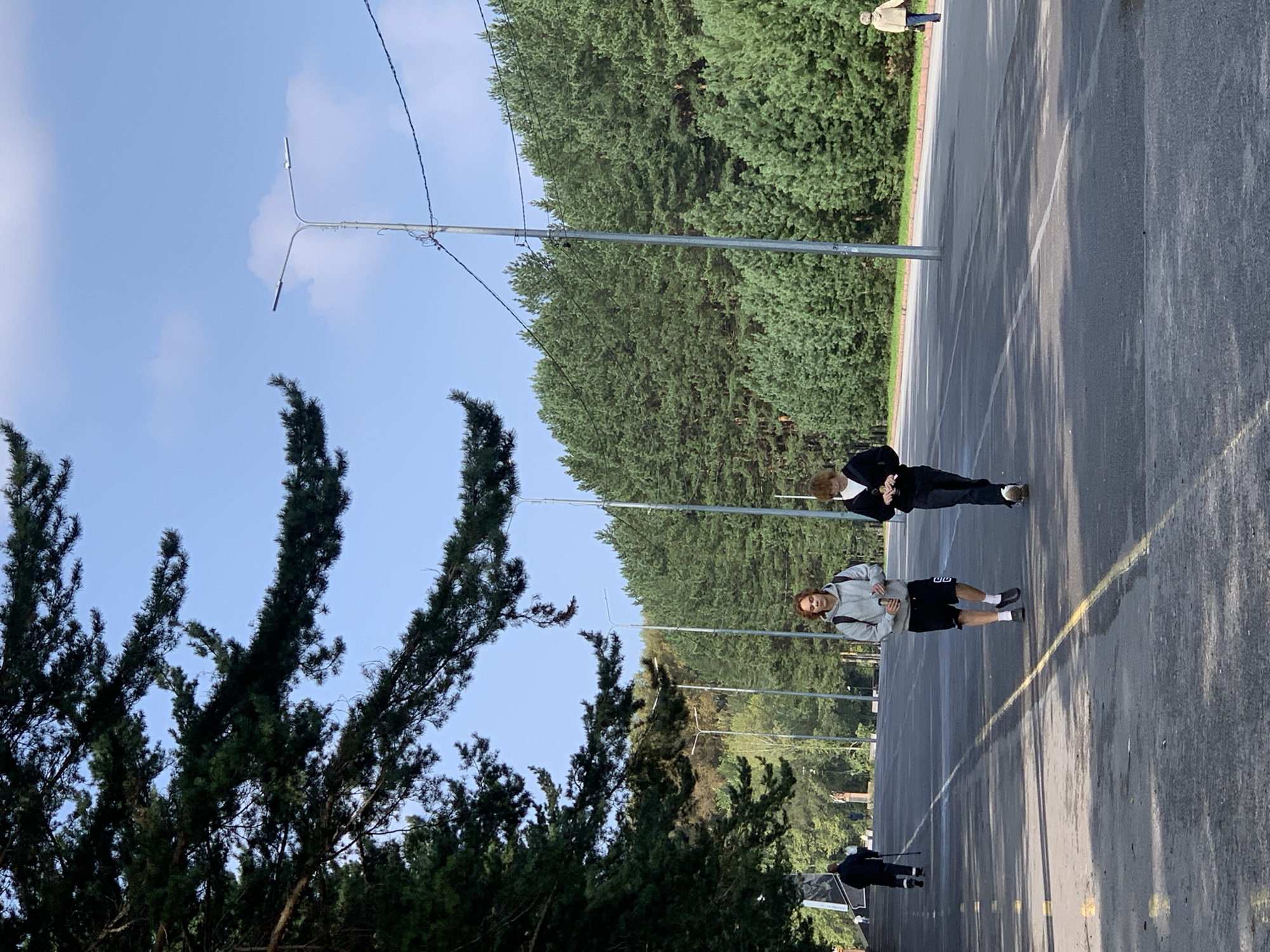 The image depicts an outdoor scene on a paved road lined with trees and streetlights. In the foreground, two individuals are walking on the road. The person on the left, wearing a grey hoodie and black shorts, is carrying a backpack and a beverage can. The person on the right, wearing a black jacket and dark pants, is also holding a beverage can. In the background, several other people are visible walking in the distance. A dense forest of evergreen trees forms the backdrop. The sky is blue with scattered clouds. This scene is located at the Victory Park in Prokopyevsk, Russia, a public space known for its monuments and recreational areas.