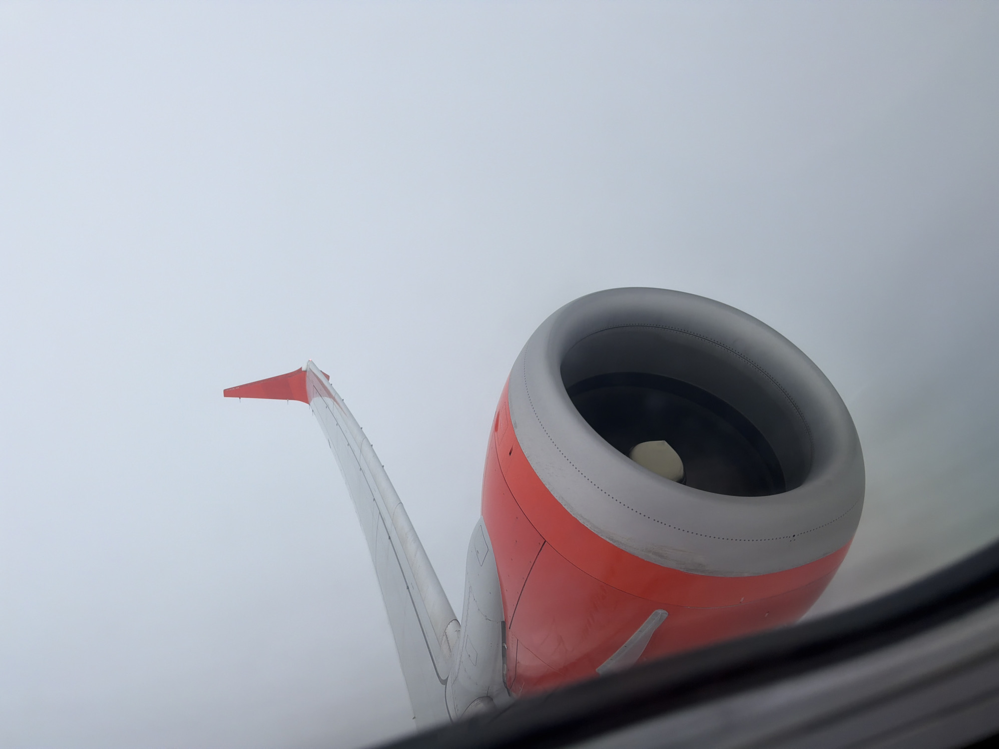 From the perspective of an airplane window, an exterior view depicts a wing with a red-tipped aileron. Adjacent to the wing is an orange engine housing with a gray inlet. The background is a uniform gray, indicative of either cloud cover or fog, suggesting the plane is in flight or just after takeoff. The location, Kuta Selatan, Indonesia, is not discernible within the visual information provided.
