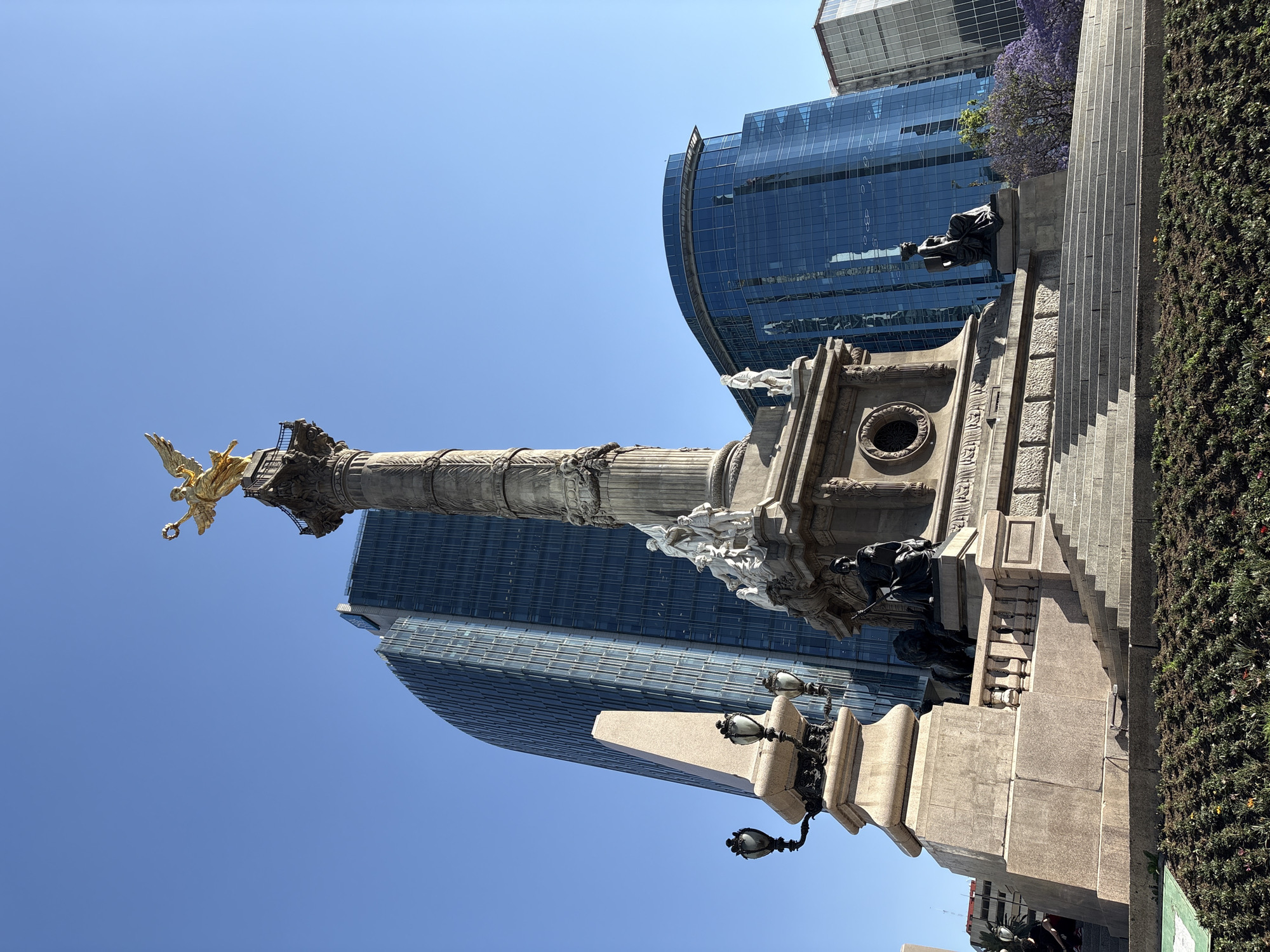 The image displays the El Ángel de la Independencia monument situated at the Glorieta del Ángel on Paseo de la Reforma in Mexico City, Mexico. The central feature is a tall, grey stone column topped by a golden, winged female figure holding a laurel wreath and a broken chain. The column rests on a large, multi-tiered stone base with steps, adorned with both white marble and dark bronze sculptures depicting allegorical and historical figures, including four seated bronze statues at the corners of the main plinth. To the left of the monument's base stands a lighter-colored stone obelisk topped with a two-lamp street fixture. In the immediate foreground, a dark green garden bed with sparse colorful flowers is visible. The background features several modern skyscrapers with reflective glass facades, one tall and slightly curved on the left, and another with a prominent rounded top on the right, under a clear blue sky. Sunlight illuminates the scene from the right, creating pronounced shadows on the monument's base and steps. Two partially visible individuals are seated on a lower stone structure to the far left, obscured by shadow.