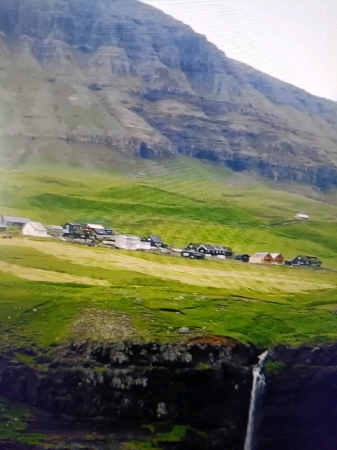 The image depicts a village nestled on a grassy hillside, with a dramatic mountain range forming the backdrop. Several houses, characterized by their dark roofs and white or wooden walls, are scattered across the landscape. A thin waterfall cascades down a rocky cliff face in the foreground. The scene is bathed in natural light, suggesting an outdoor setting. There are no people visible in the image, nor any indication of activity. The geographical features suggest a remote, possibly coastal or mountainous region, and there is no connection to the city of Netanya, Israel.