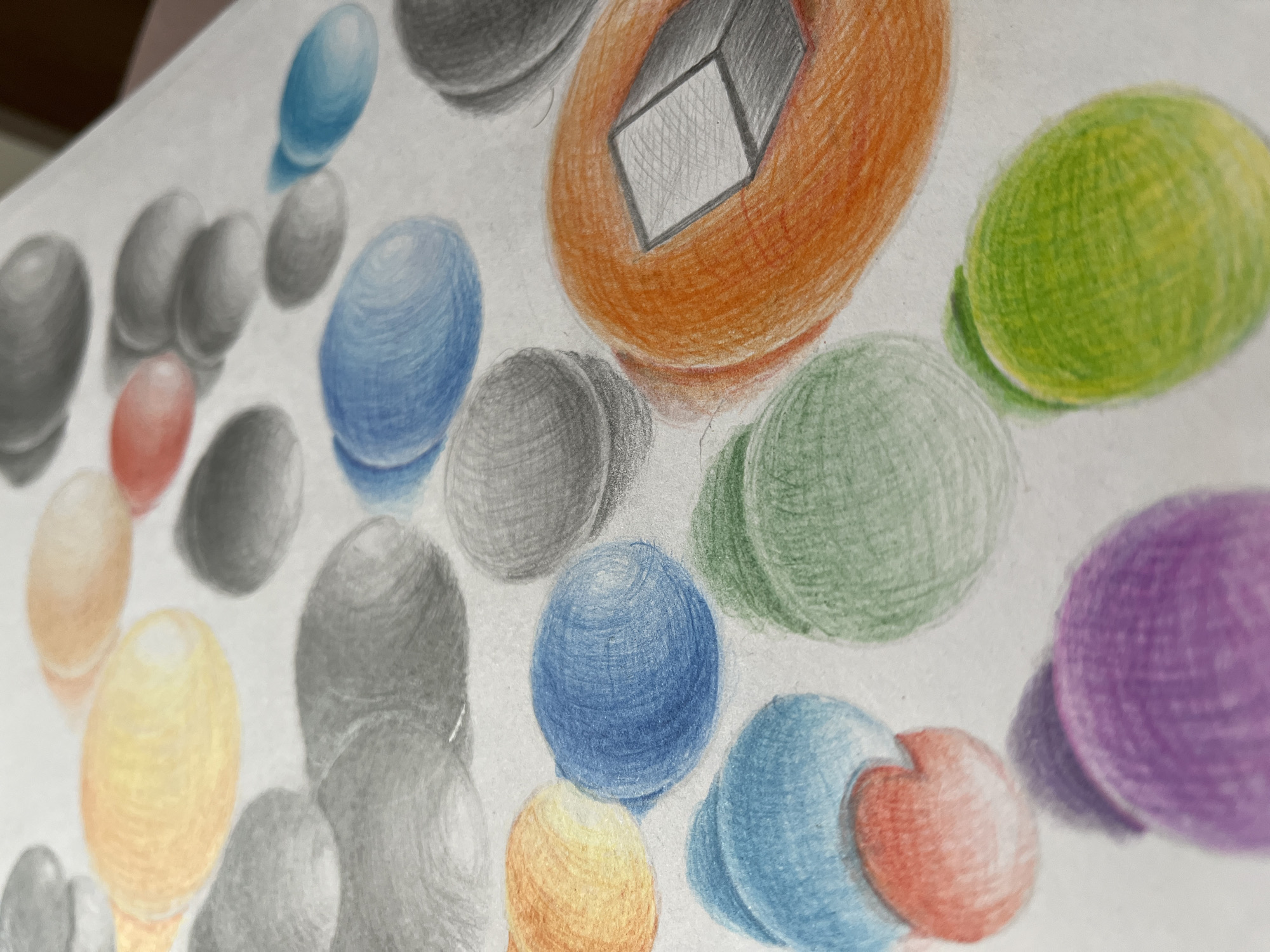 The image depicts a close-up, overhead view of a collection of colorful, smooth, oval-shaped objects, likely rendered in colored pencil on white paper. These objects are arranged in a scattered pattern across the surface, with some overlapping. The colors present include yellow, orange, red, blue, green, gray, and purple. The rendering style emphasizes the roundness and texture of the objects, with visible pencil strokes creating subtle shading and highlights. Some of the objects appear to cast soft shadows. There is no indication of a specific location, people, or actions occurring. The image is a still life drawing of stones or marbles. The city of Krasnyye Barrikady, Russia is not visible or implied in this image.