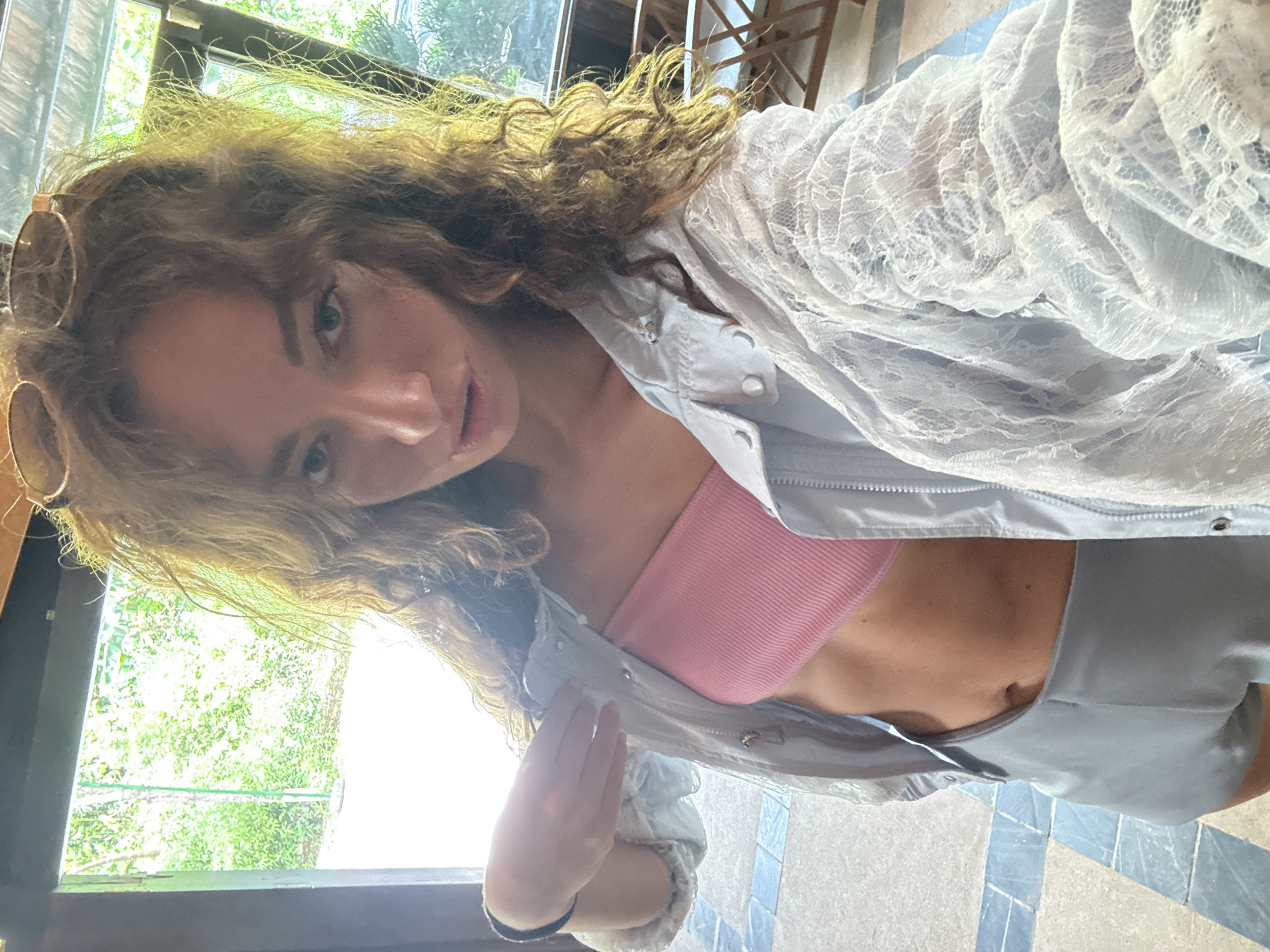 A female-presenting individual is captured in a front-facing selfie, positioned indoors. Her long, light brown to auburn curly hair frames her face and shoulders, with sunglasses resting atop her head. She gazes directly at the camera with her lips slightly parted. She wears a ribbed pink bandeau top, exposing her midriff and navel, paired with light grey shorts. An open light blue jacket, featuring a subtle lace texture on its lower sections and sleeves, is worn over the top. Her left hand, adorned with a black band on the wrist, is raised near her left shoulder. The individual stands on a tiled floor patterned with repeating light and dark blue/grey geometric squares. Behind her, large windows or an open doorway reveal a brightly lit outdoor scene dominated by dense green foliage. To the right, through another window, light-colored wooden chairs and tables are partially visible, suggesting an outdoor seating area. The image provides no visual information to ascertain a specific city or country location such as Bản Đêu, Vietnam.