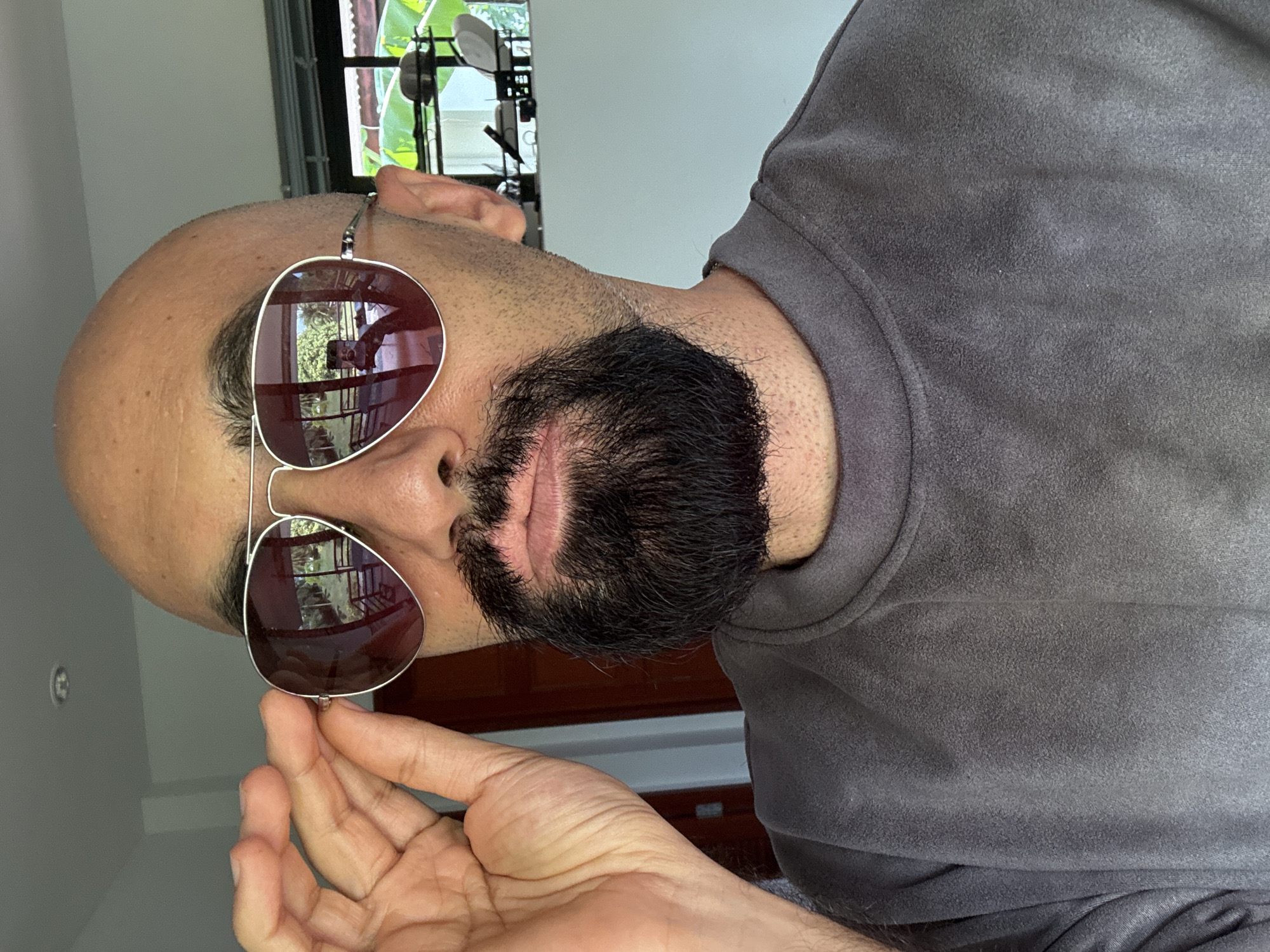 A bald man with a goatee and sunglasses is adjusting his glasses with his left hand. He is wearing a gray sweatshirt. The background appears to be an indoor setting with a window that shows greenery and some outdoor structures. There are reflections of the surroundings in the sunglasses, including what appears to be a camera or phone being held to take the picture. The man is looking directly at the camera.