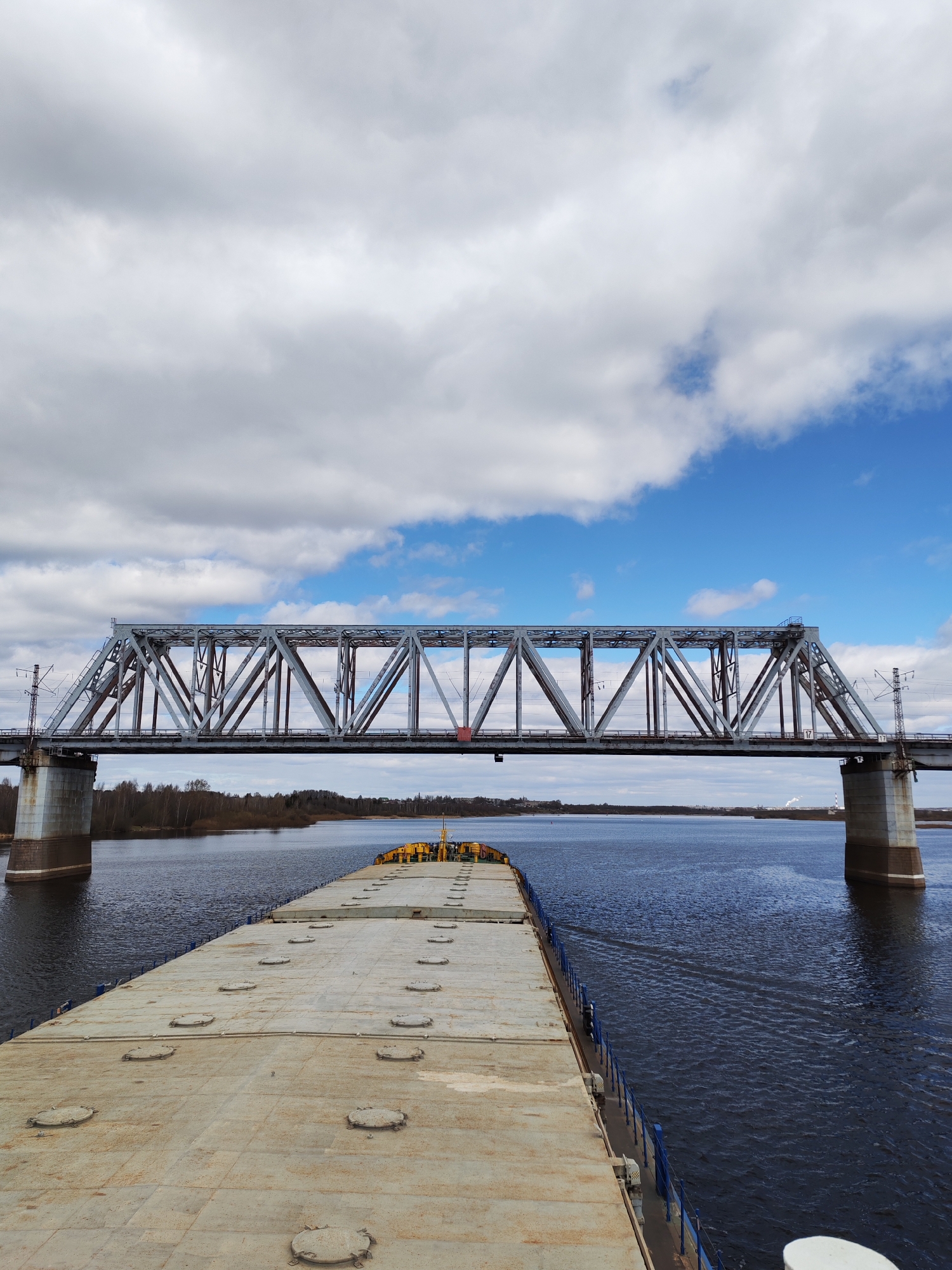 The image shows a wide-angle view of a large flat-top barge navigating a wide river, with a steel truss bridge spanning overhead. The barge's deck is characterized by a series of circular access covers. In the background, a tree line is visible on the riverbank, and a distant industrial facility with smokestacks is present on the right. The sky is partially cloudy, with patches of blue interspersed among the white clouds. The bridge is constructed with steel girders and rests on concrete pylons. The location is the city of Unknown, Unknown.